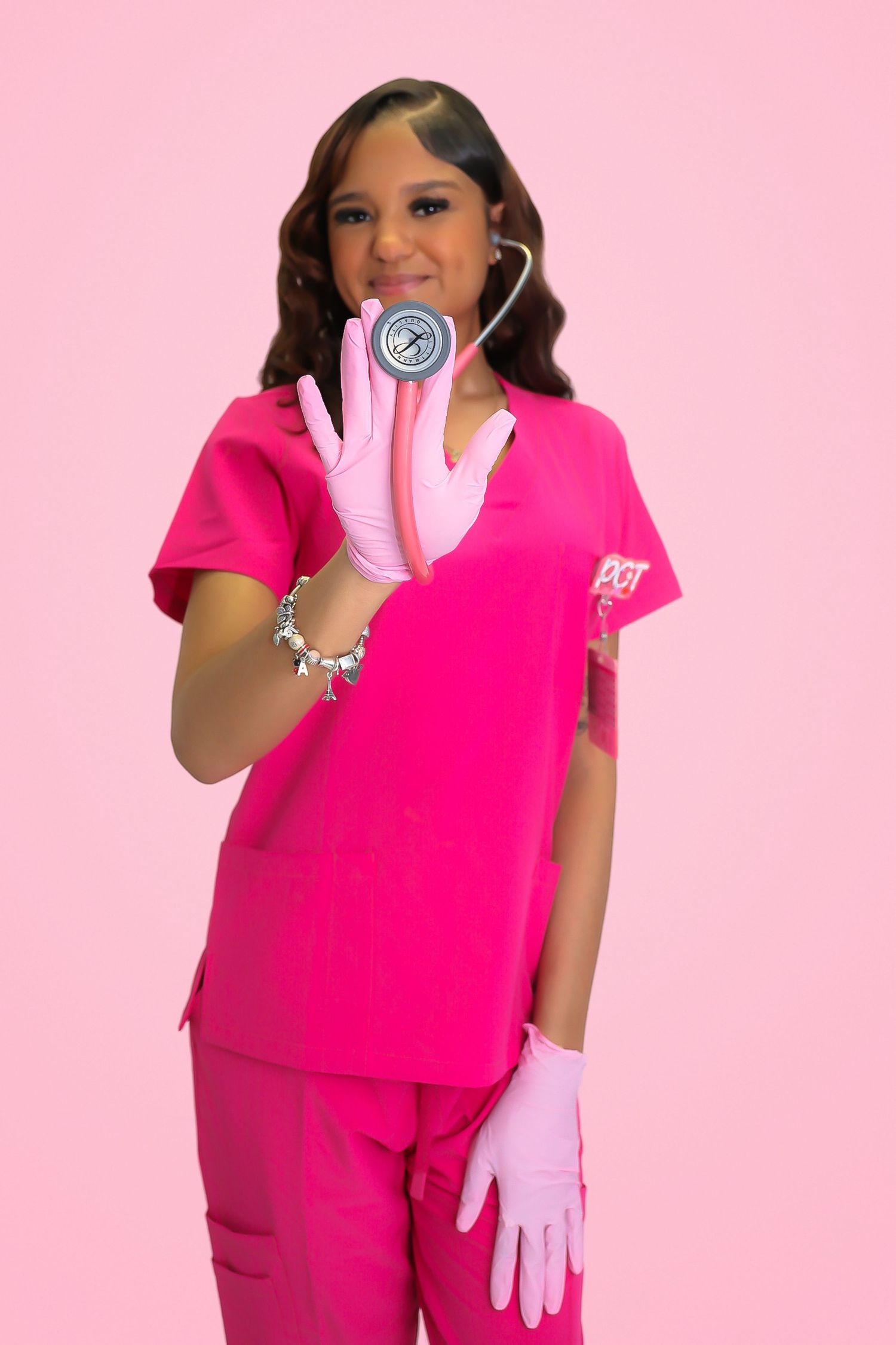 Young female nurse in pink scrubs and gloves holds stethoscope against pink background.