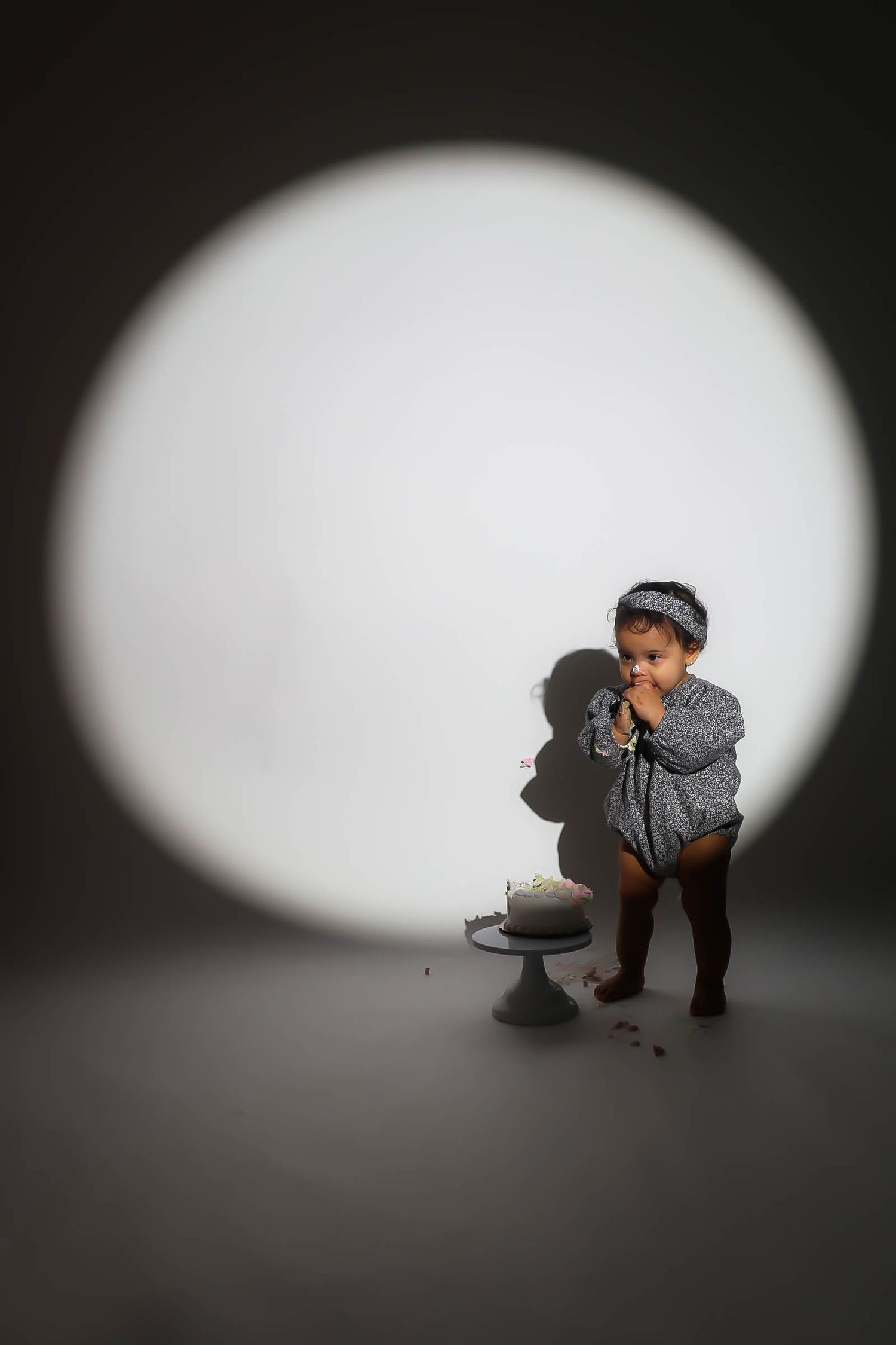 A toddler in a gray romper eats birthday cake in a dramatic spotlight during a first birthday cake smash photo session.