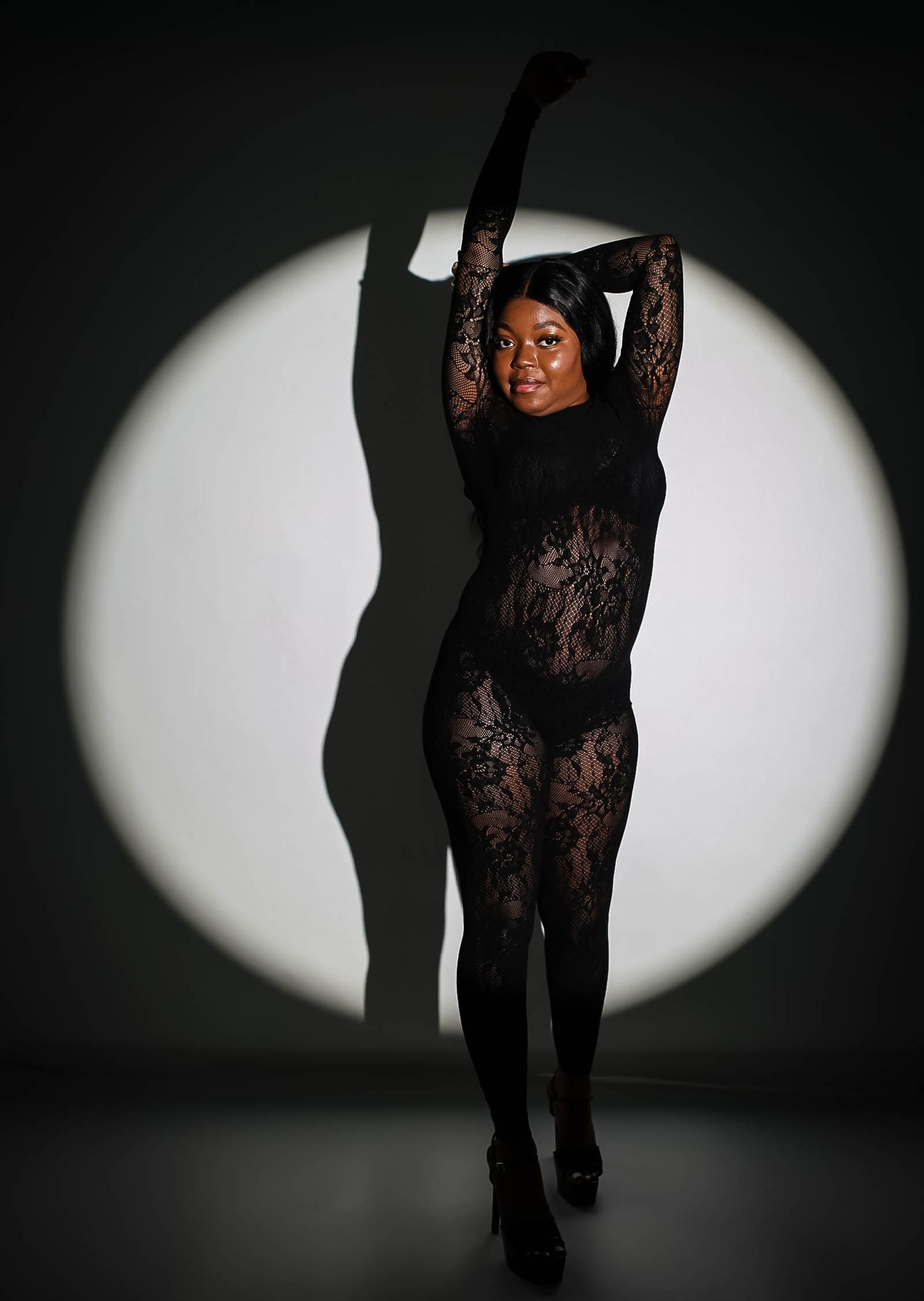 Black woman in sheer black lace bodysuit posing with arms raised in dramatic spotlight against dark background.