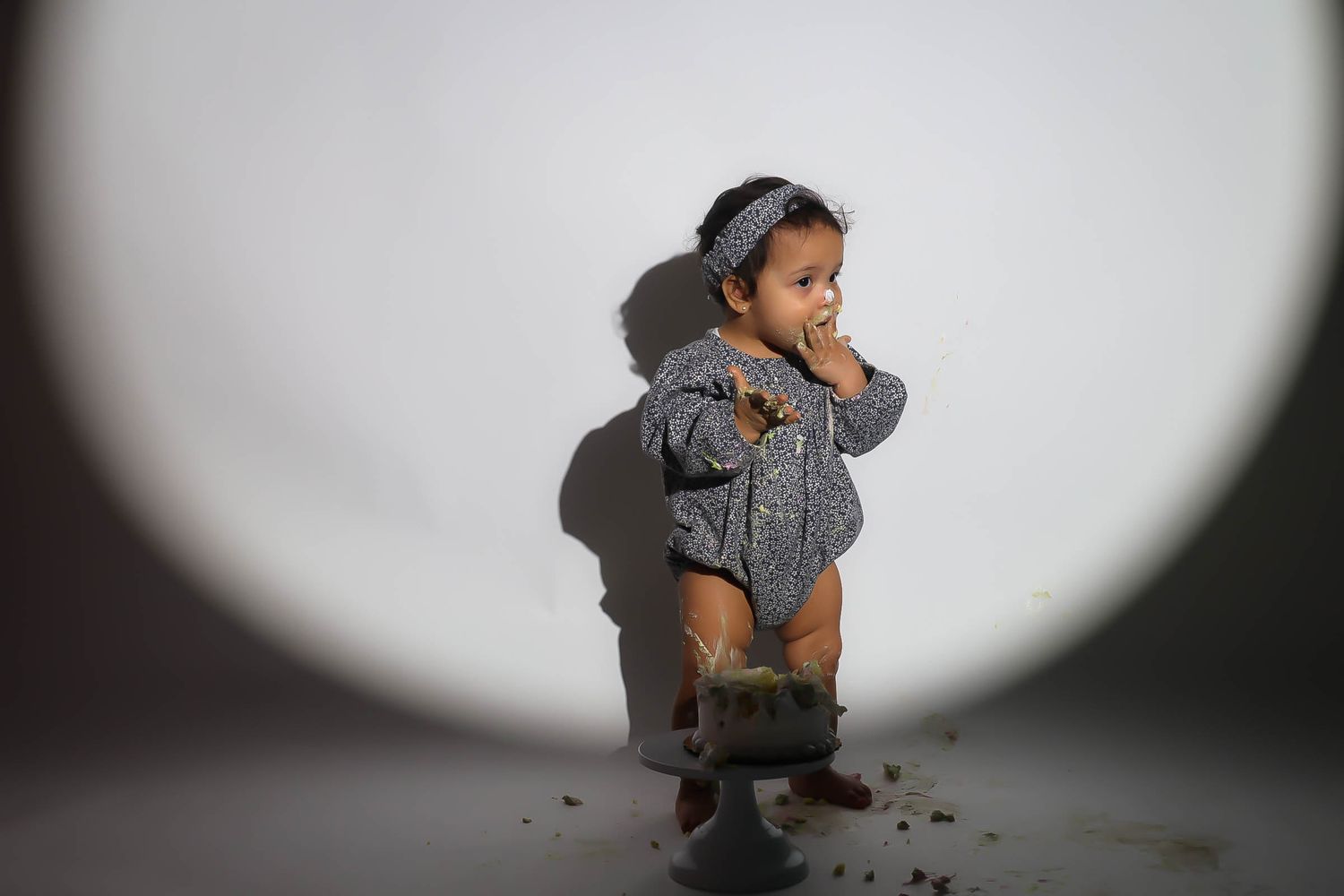 Adorable baby girl in gray romper and headband standing on a cake smash set with white background.