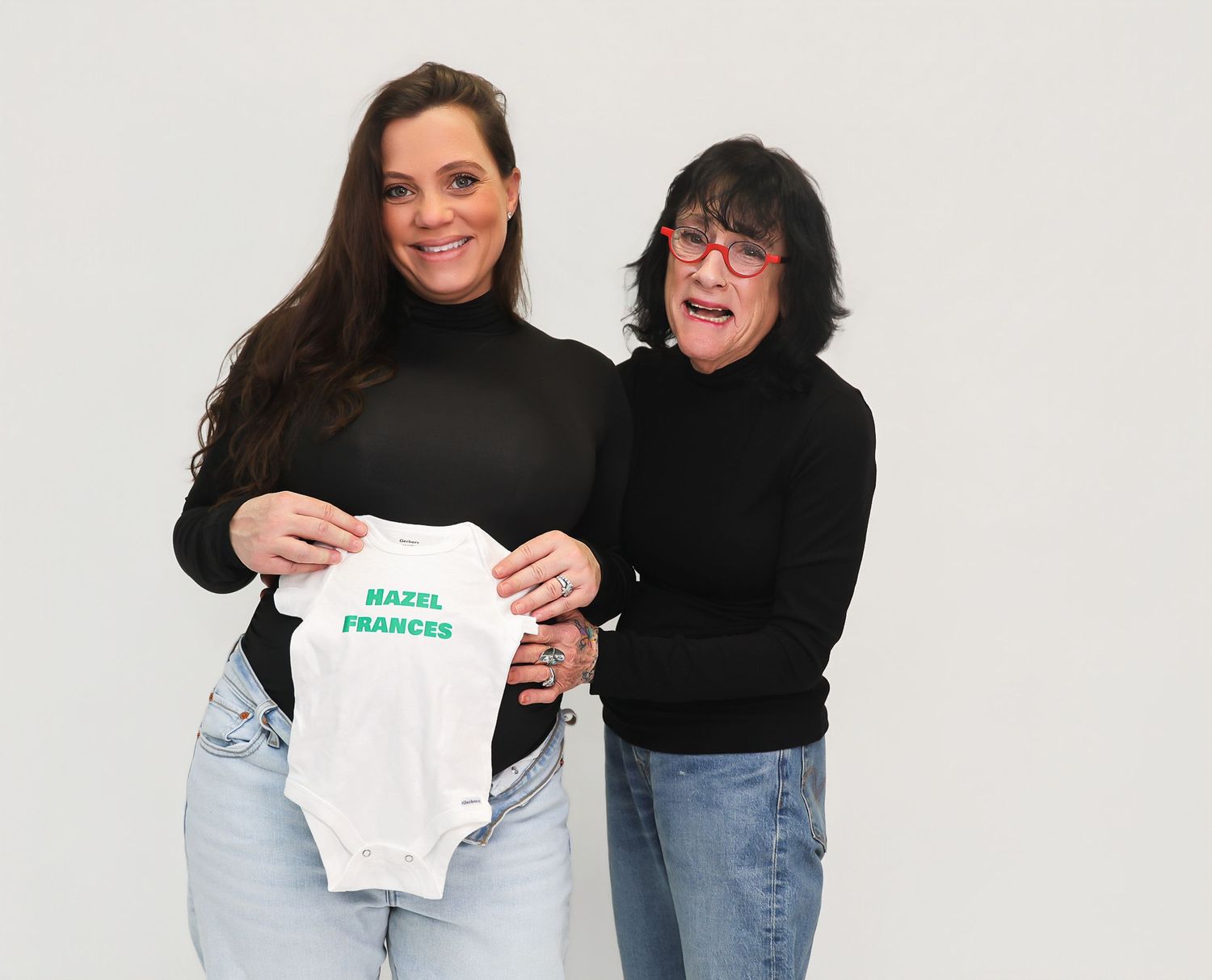 Two women smiling and holding a white baby onesie printed with the name Hazel Frances against a white background.