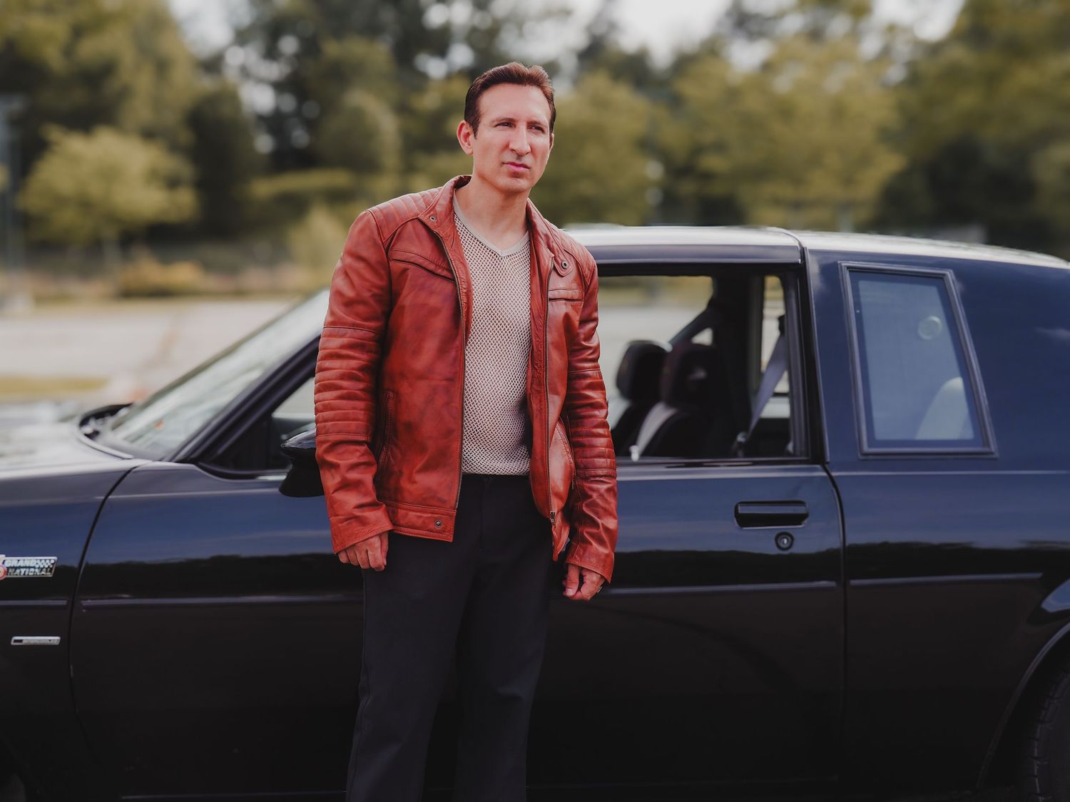 Person in red leather jacket stands next to a black car in an outdoor setting.