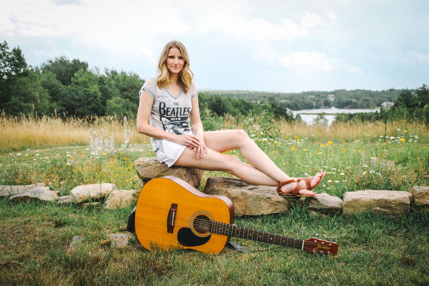 A person sits on a log in a wildflower meadow with an acoustic guitar and lake view in the background.