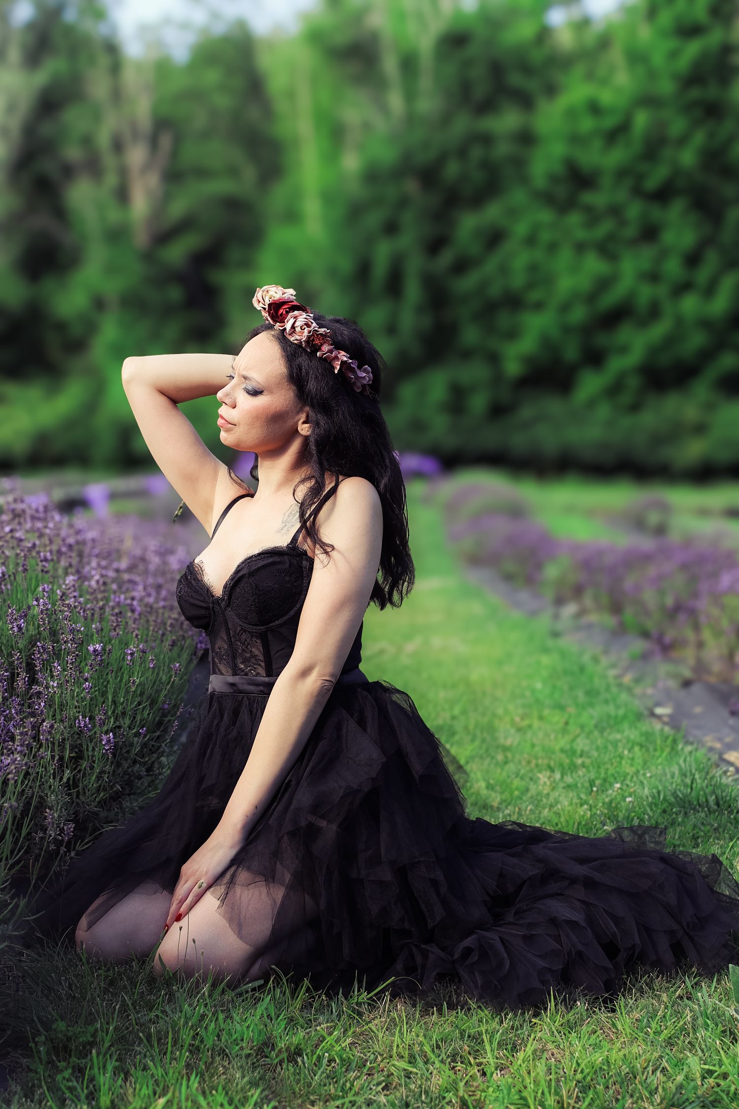 A figure in a flowing black dress poses gracefully among blooming lavender flowers in a lush garden setting.