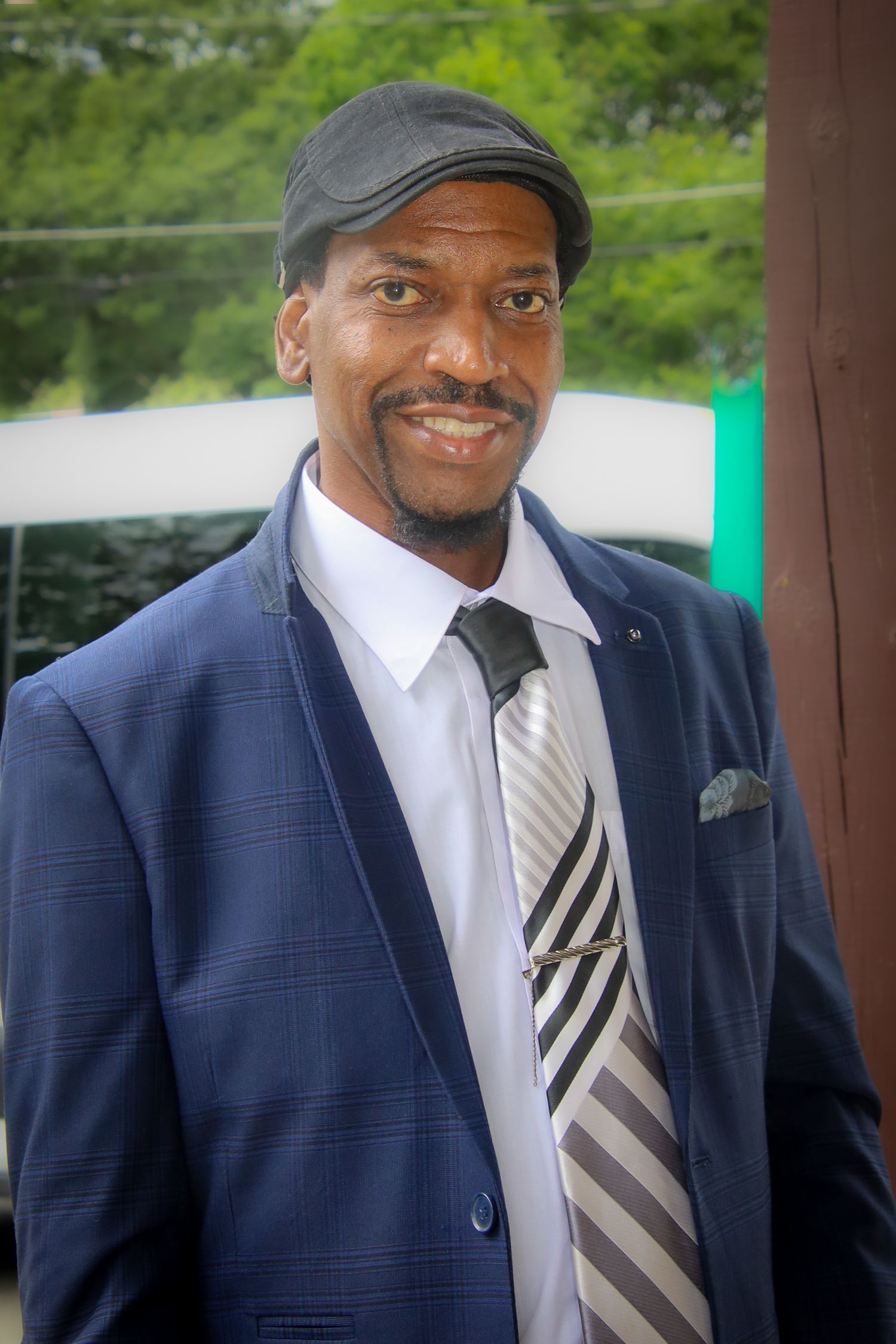 A person in a navy pinstripe suit and striped tie smiles warmly while wearing a dark cap.