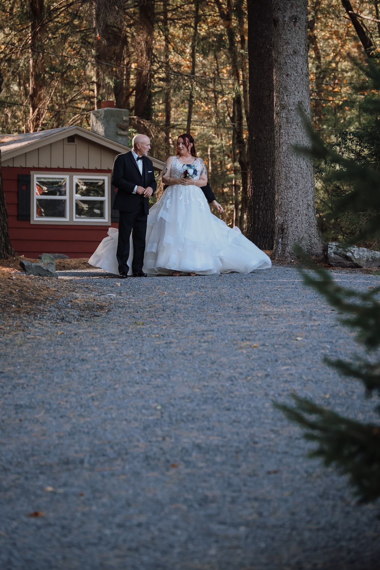 A couple in formal wedding attire stands together on a gravel path near a rustic cabin in the woods.