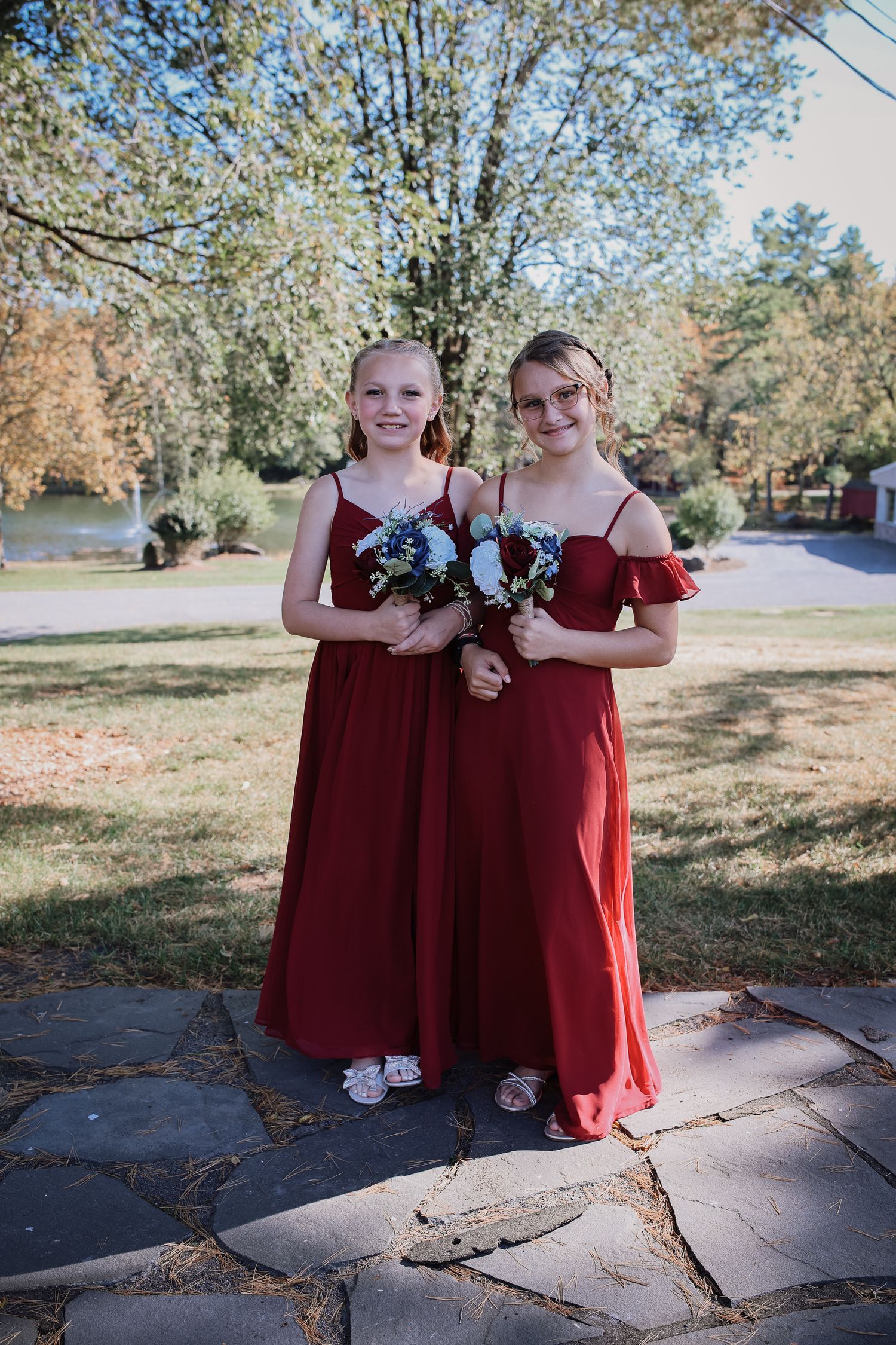 Two people in burgundy formal dresses stand together on stone pavement holding floral bouquets in an outdoor setting.