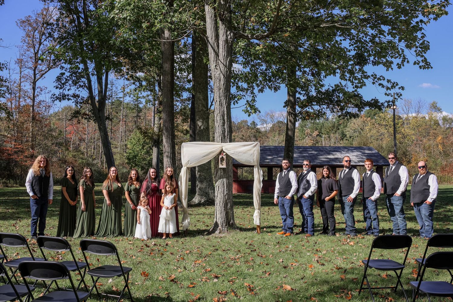 A wedding party poses outdoors in autumn with trees and blue chairs arranged on a grassy lawn.