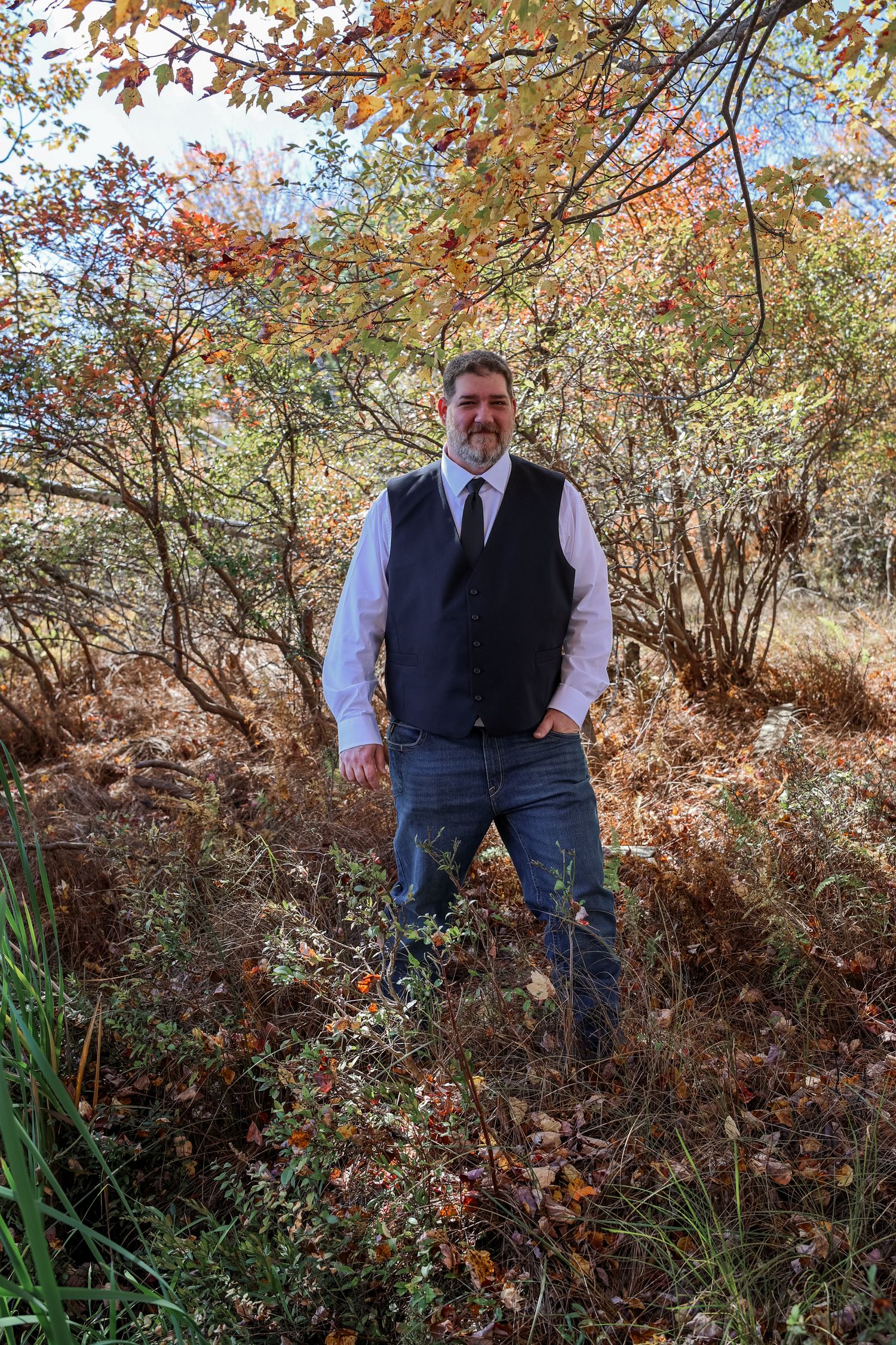 Person in navy vest and white shirt stands among autumn trees and bushes with changing fall foliage colors.