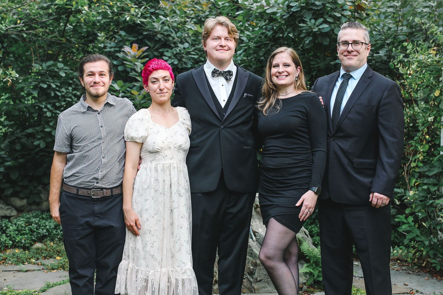 A group of formally dressed people pose together outdoors in front of green foliage.
