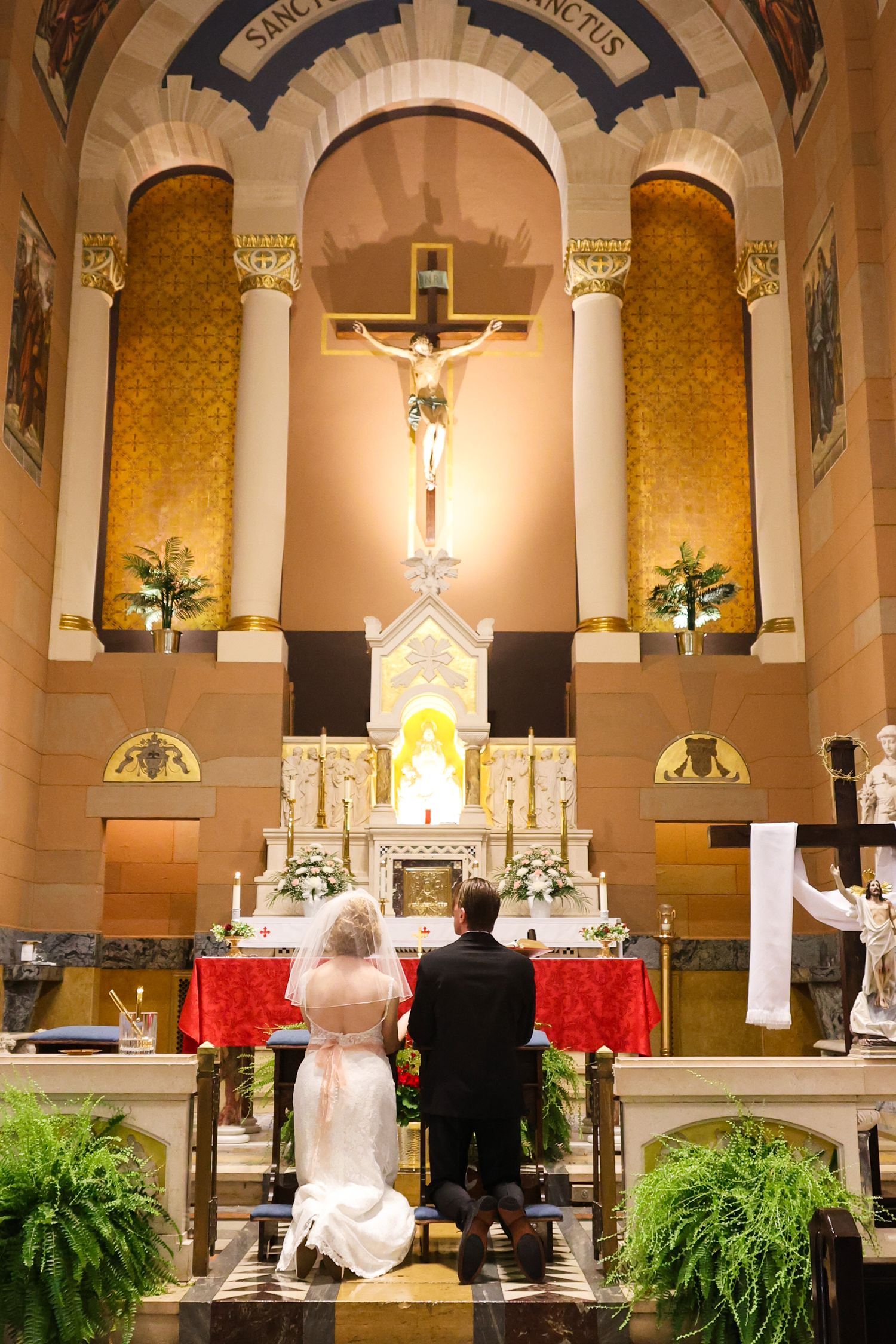 Interior of a Catholic church during a wedding ceremony with ornate altars, crosses and decorative plants.