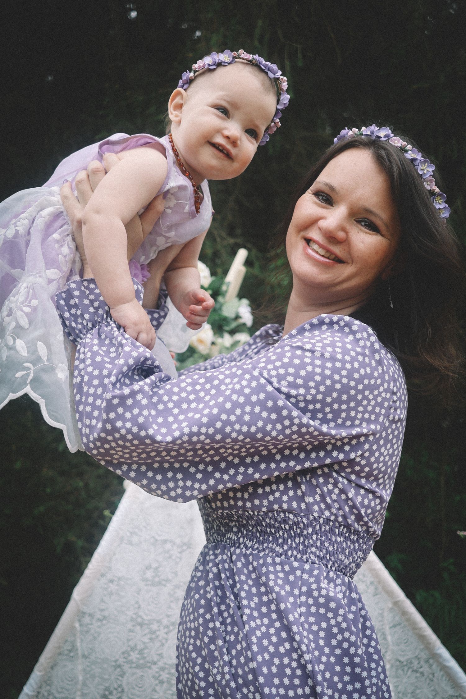 A woman in a polka dot dress holds and plays with a baby wearing white lace in an outdoor setting.