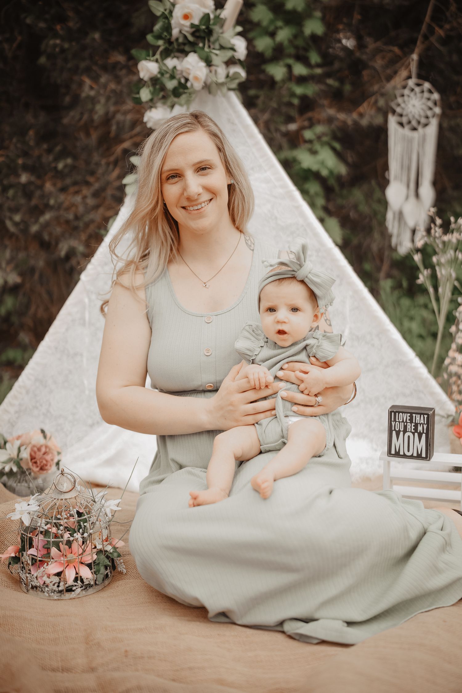 A mother in a sage green dress sits with her baby in an outdoor garden setting with floral decorations and rustic decor.