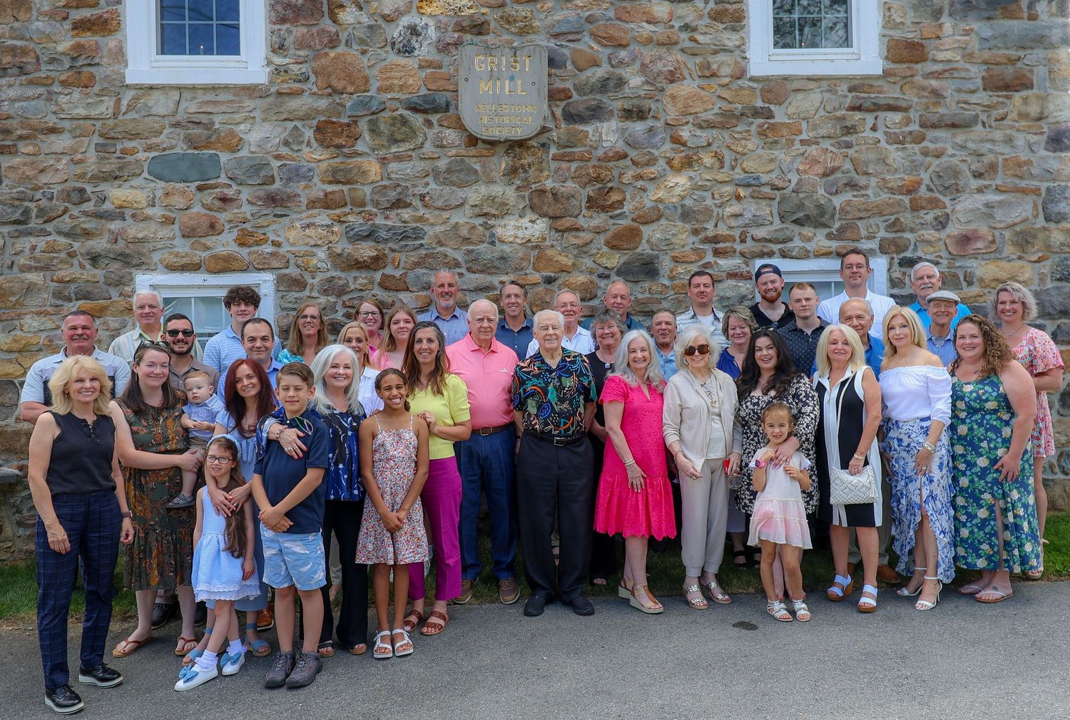 A large group of people gathered for a formal occasion against a rustic stone wall exterior.