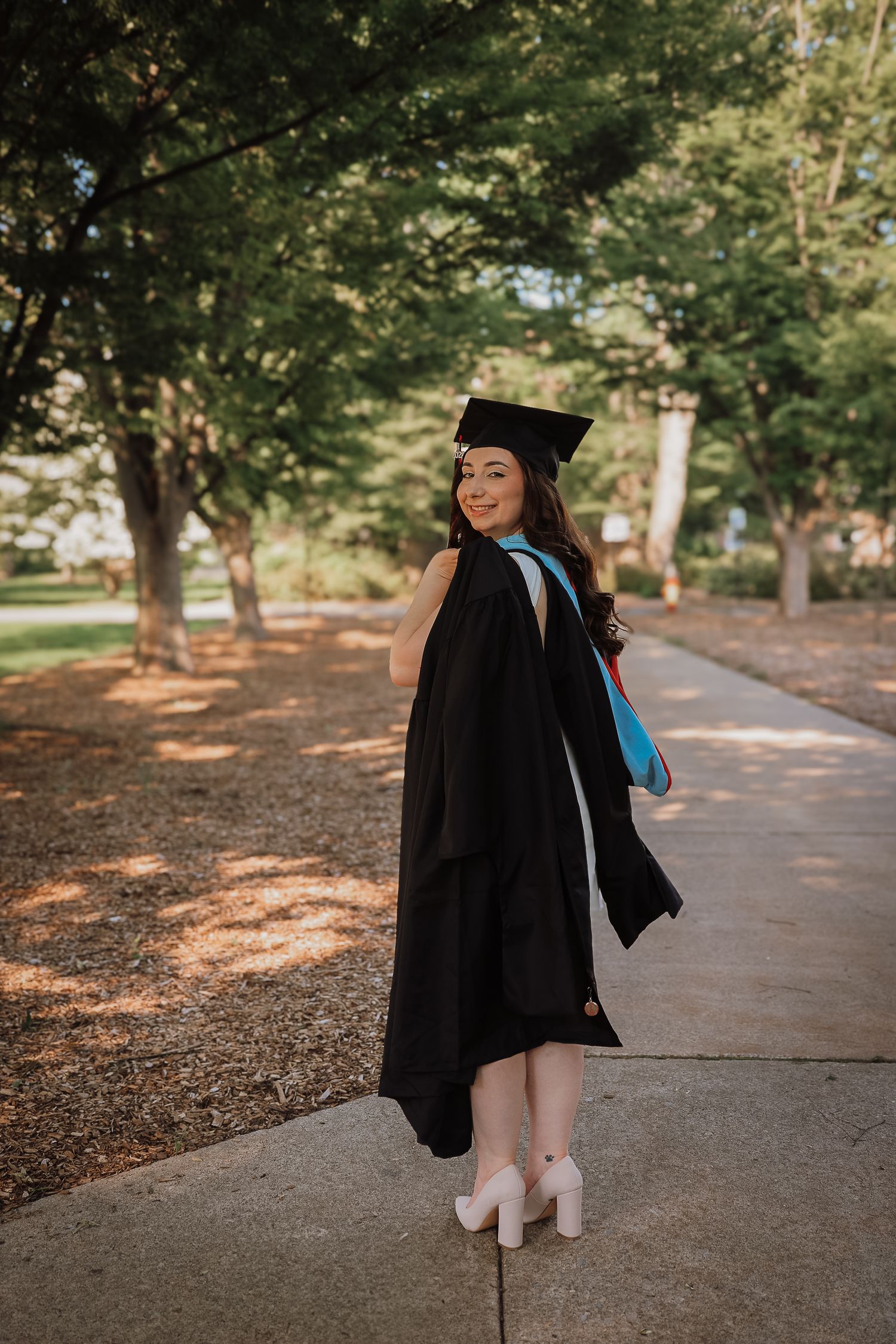 Graduate in black cap and gown stands on campus pathway in dappled sunlight during graduation photoshoot.