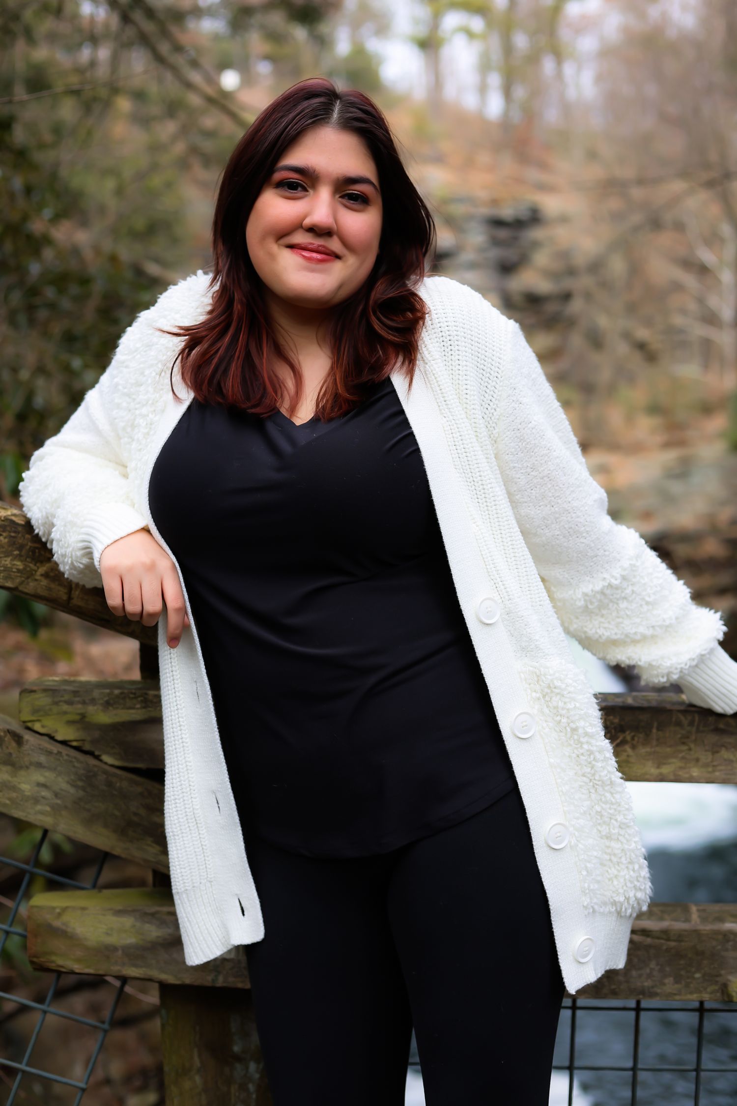 A person wearing a black top and white cardigan poses outdoors against a rustic wooden fence in autumn.