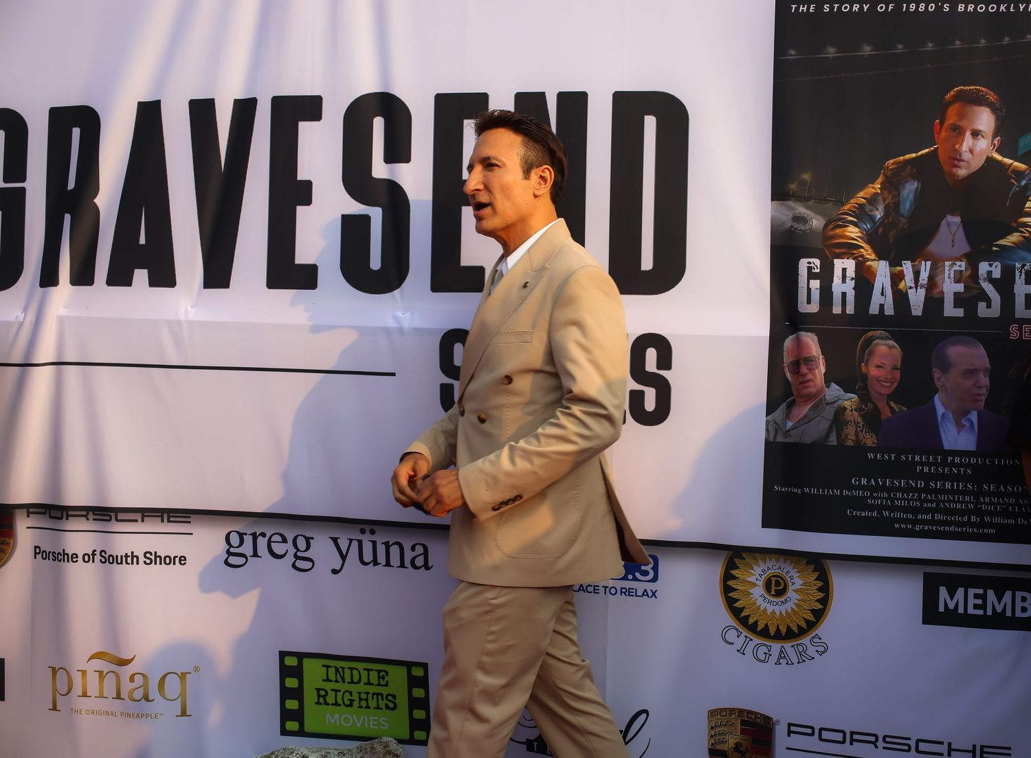 A person in a beige suit poses at a movie premiere event for 'Gravesend' with promotional posters in the background.