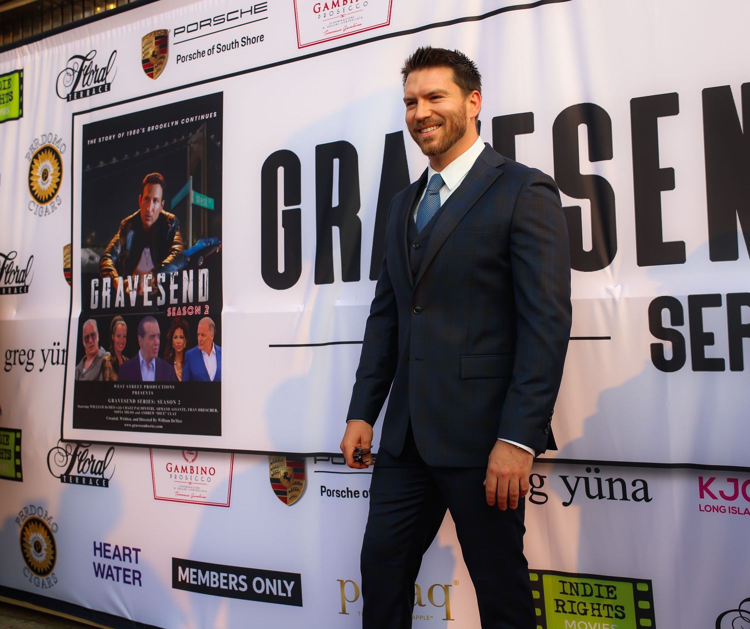 People pose for photos on a red carpet event with branded backdrop displaying logos and text.