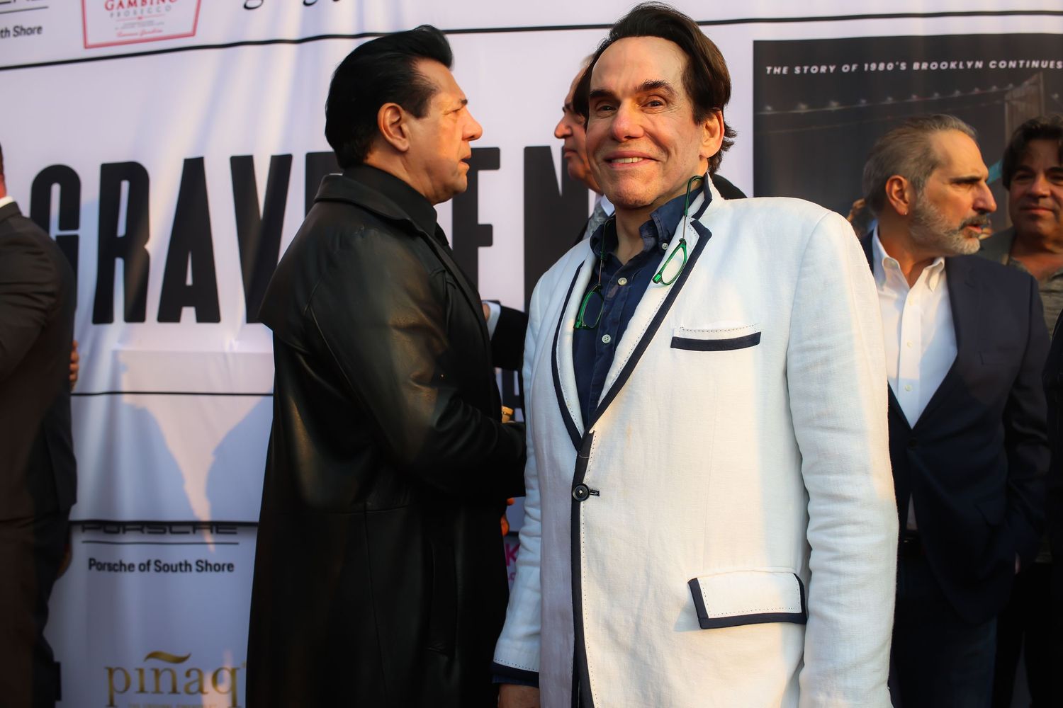 Two people in formal attire stand together at an event, one wearing a white jacket with black trim.