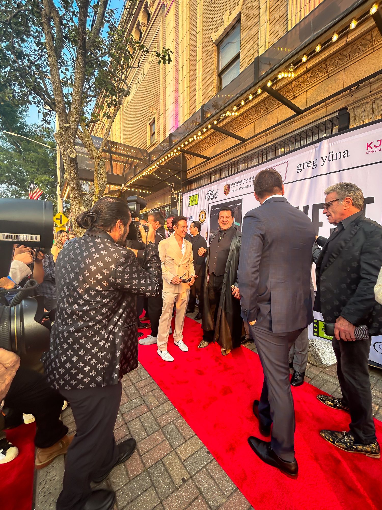 Photographers and attendees gather on a red carpet outside a building during an evening event.