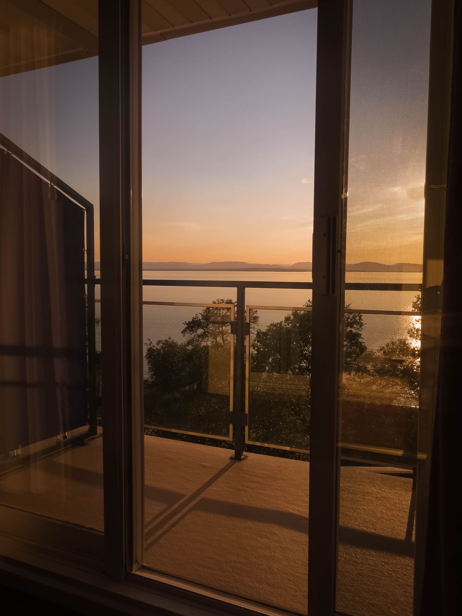Sunset view through glass balcony doors overlooking tranquil water and distant islands.
