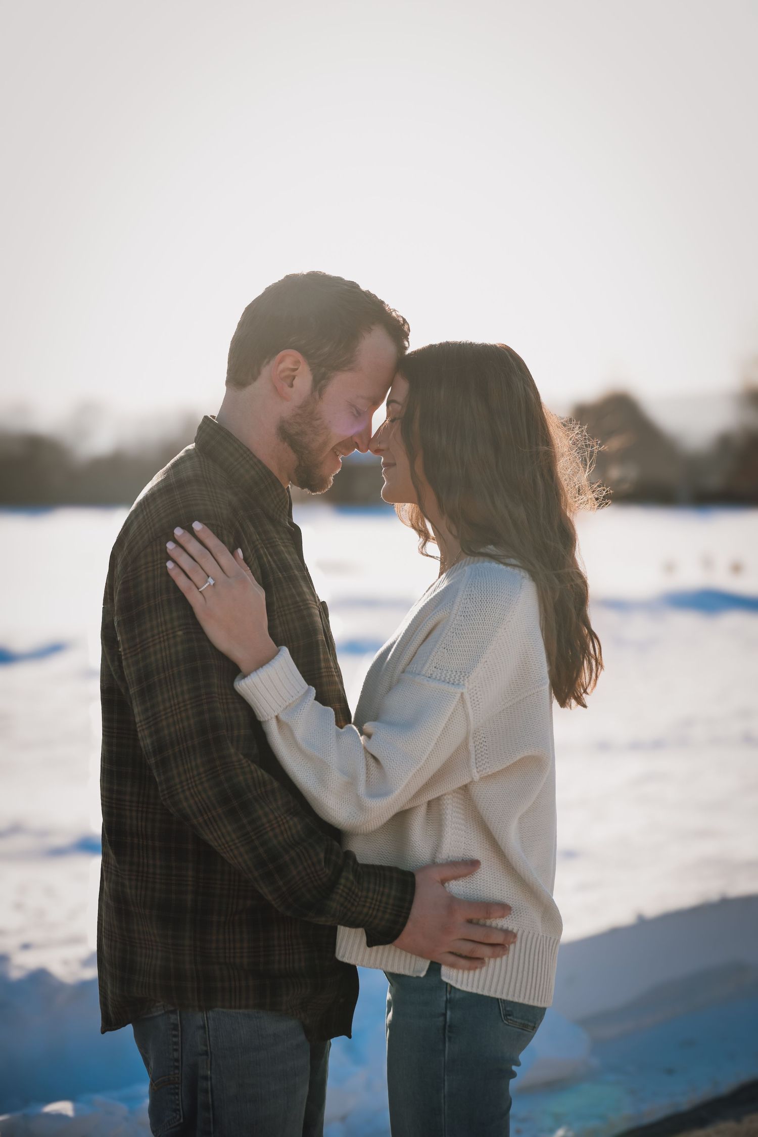 A romantic winter engagement photo of a couple embracing in the snow during golden hour sunset.