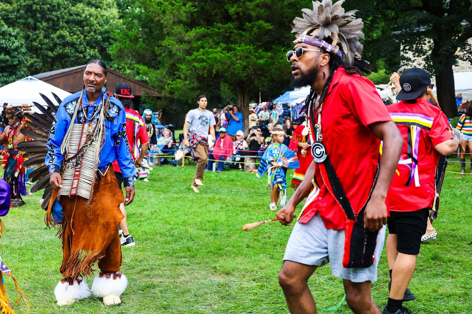 Native American dancers in traditional regalia perform at an outdoor powwow celebration in a grassy field.