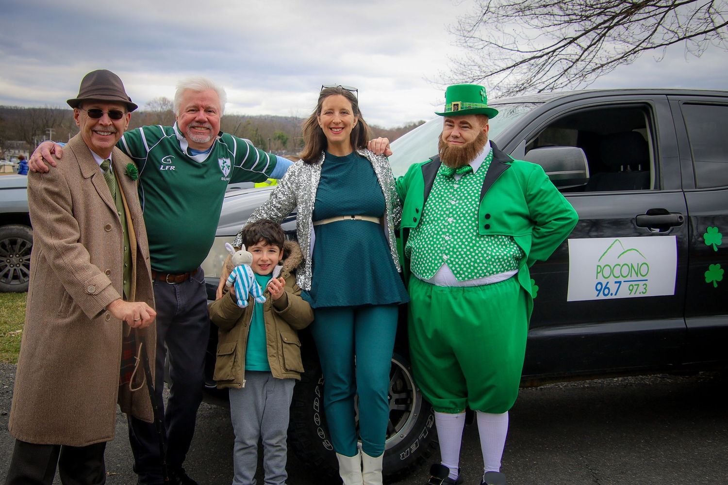A group of people wearing green clothing and festive attire gather near vehicles during a St. Patrick's Day celebration.