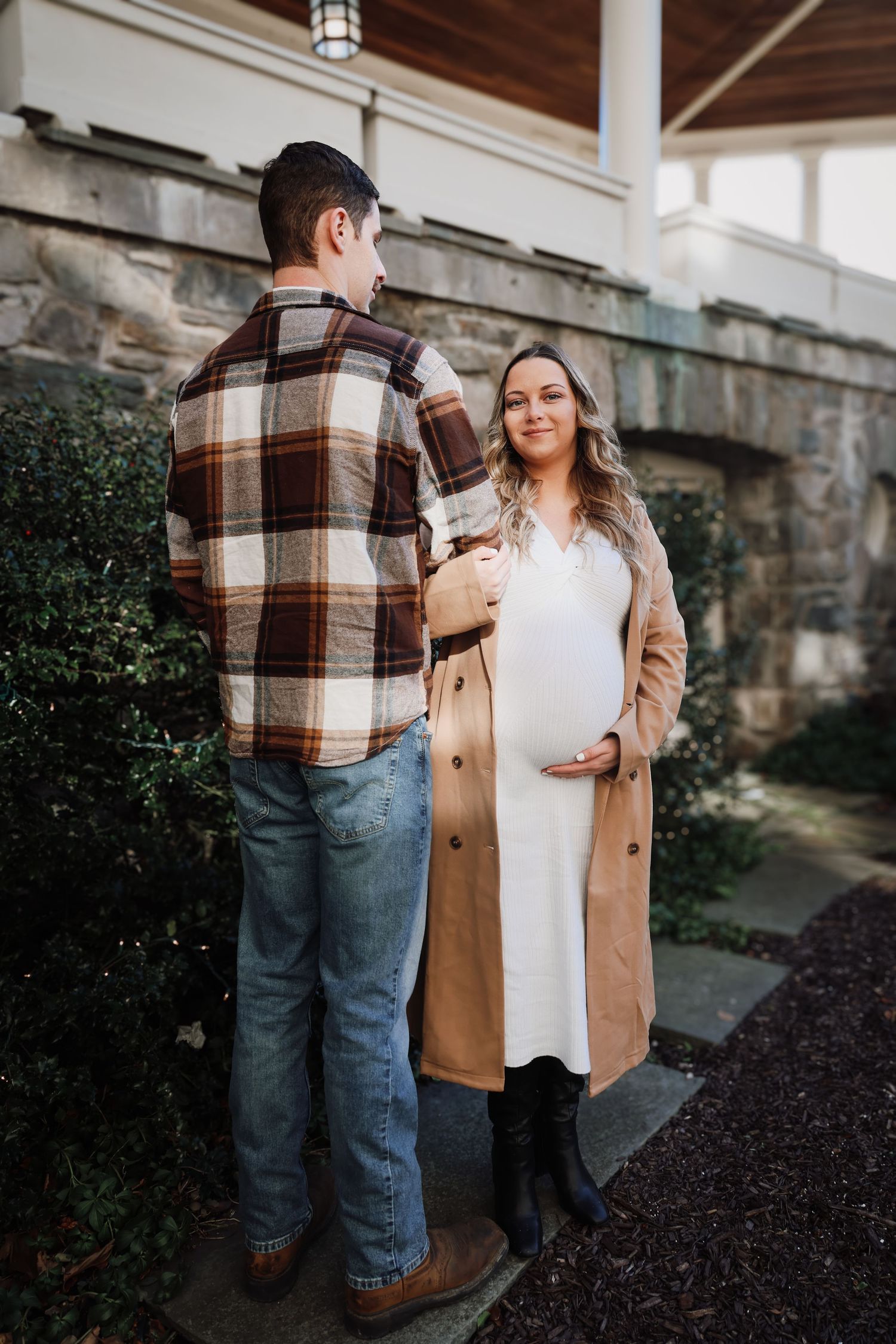 Expectant couple shares a tender moment outdoors, with her in a tan coat and him wearing a plaid flannel shirt.