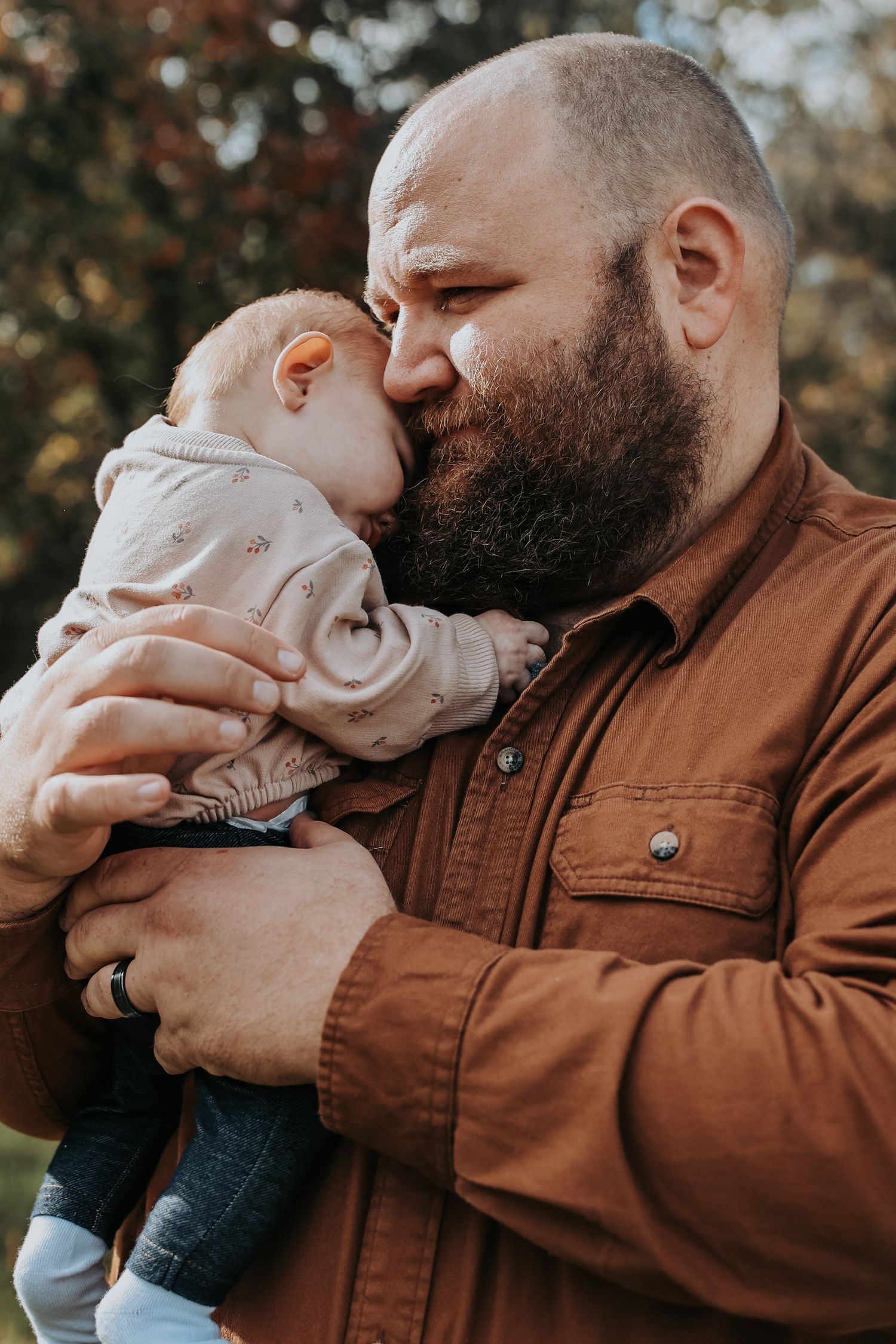 A tender moment captured outdoors as a bearded adult in a brown jacket holds and snuggles with a small baby in light clothing.
