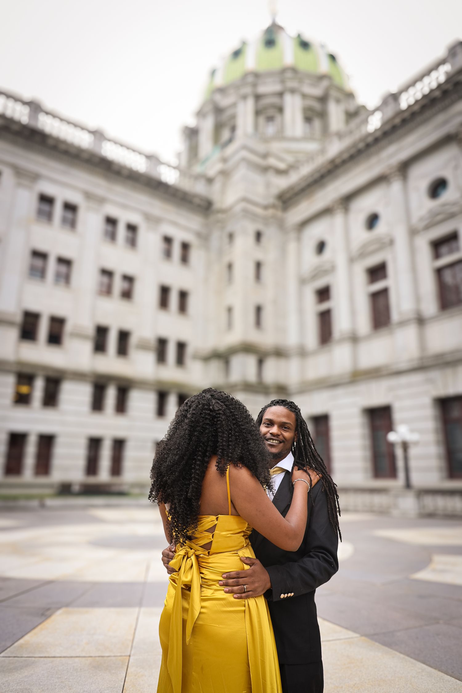 A couple in formal attire embraces on the steps of a grand government building with ornate architecture.
