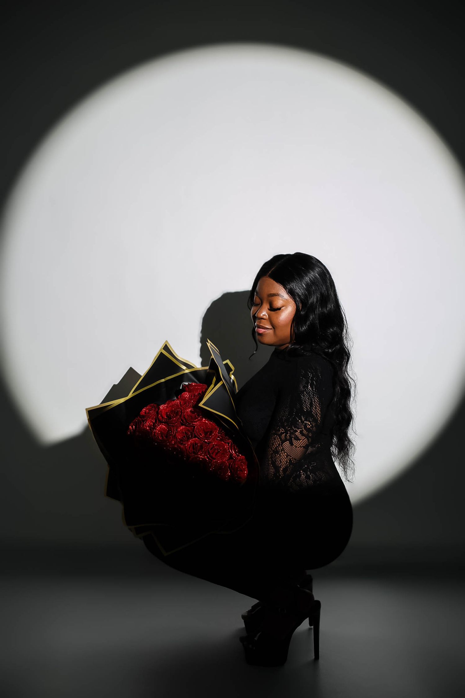 Woman in black lace outfit crouching while holding a bouquet of red roses against a spotlight background.