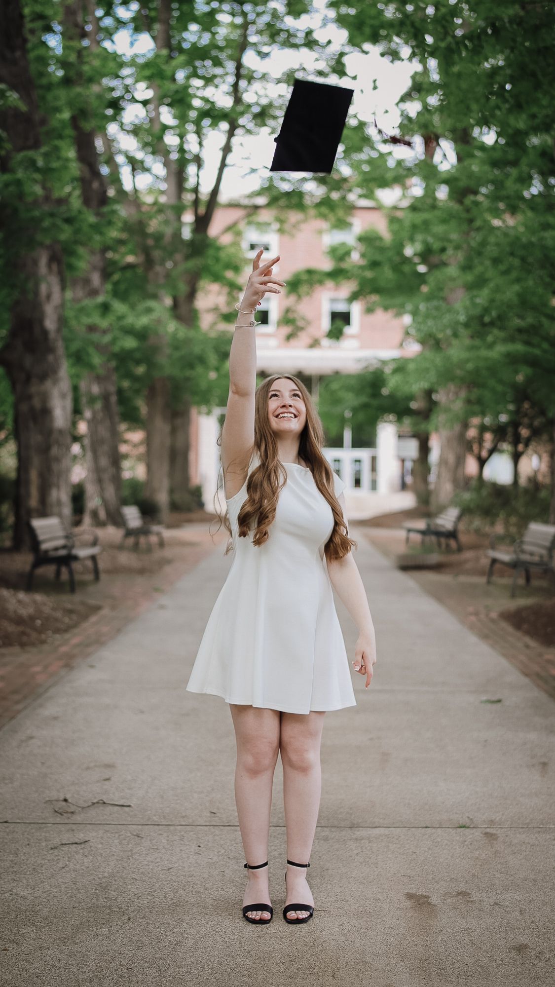 Young woman in white dress tossing graduation cap in tree-lined campus walkway, celebrating academic achievement.