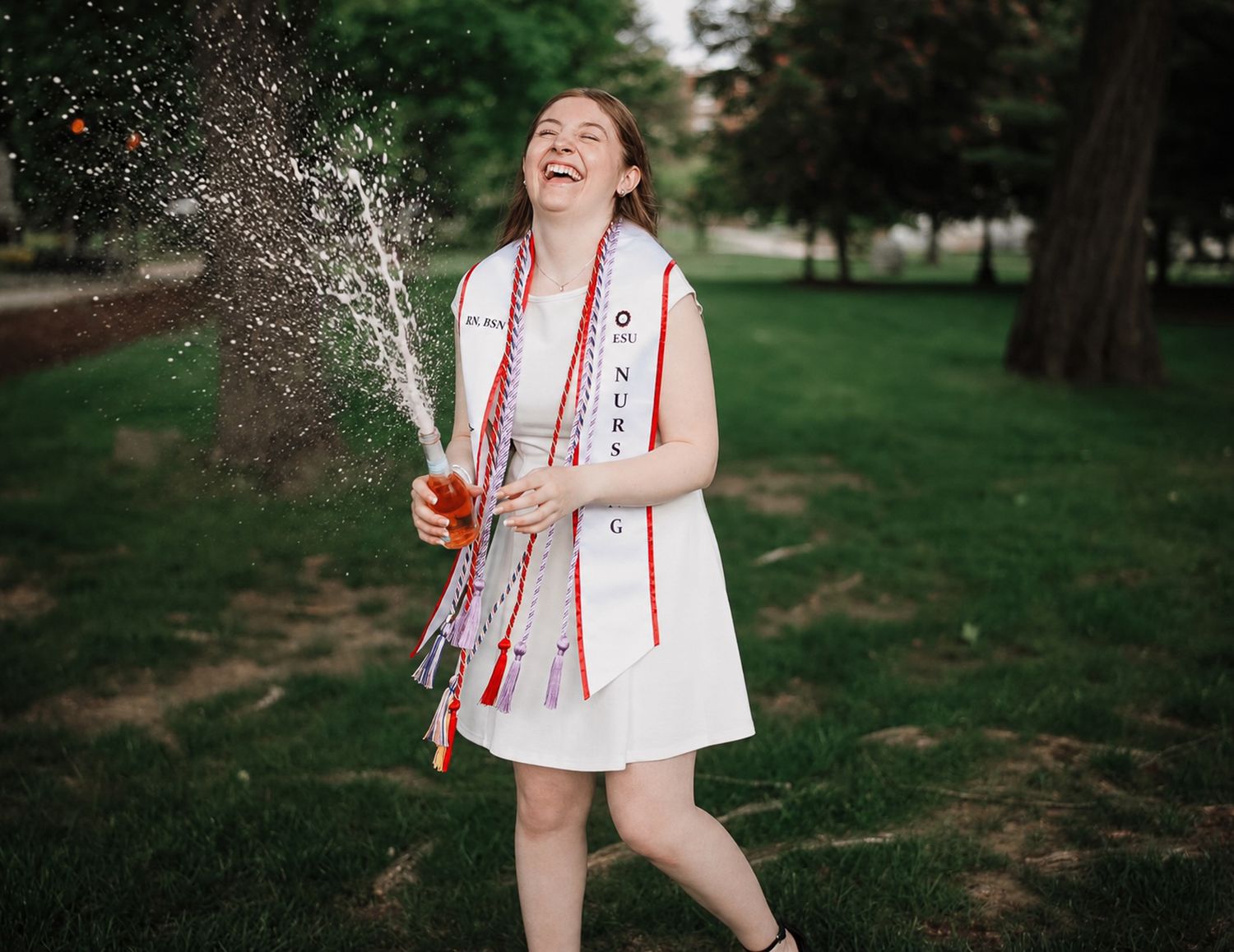 Joyful graduate in white dress and red sash celebrates outdoors, popping champagne bottle on green lawn.