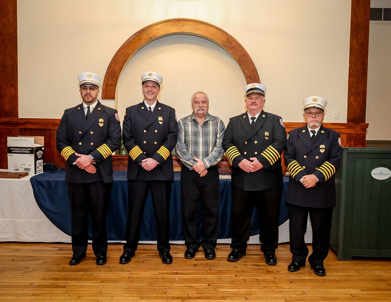 Five firefighters in dress uniforms pose for a formal group photo inside a banquet hall with an arched backdrop.