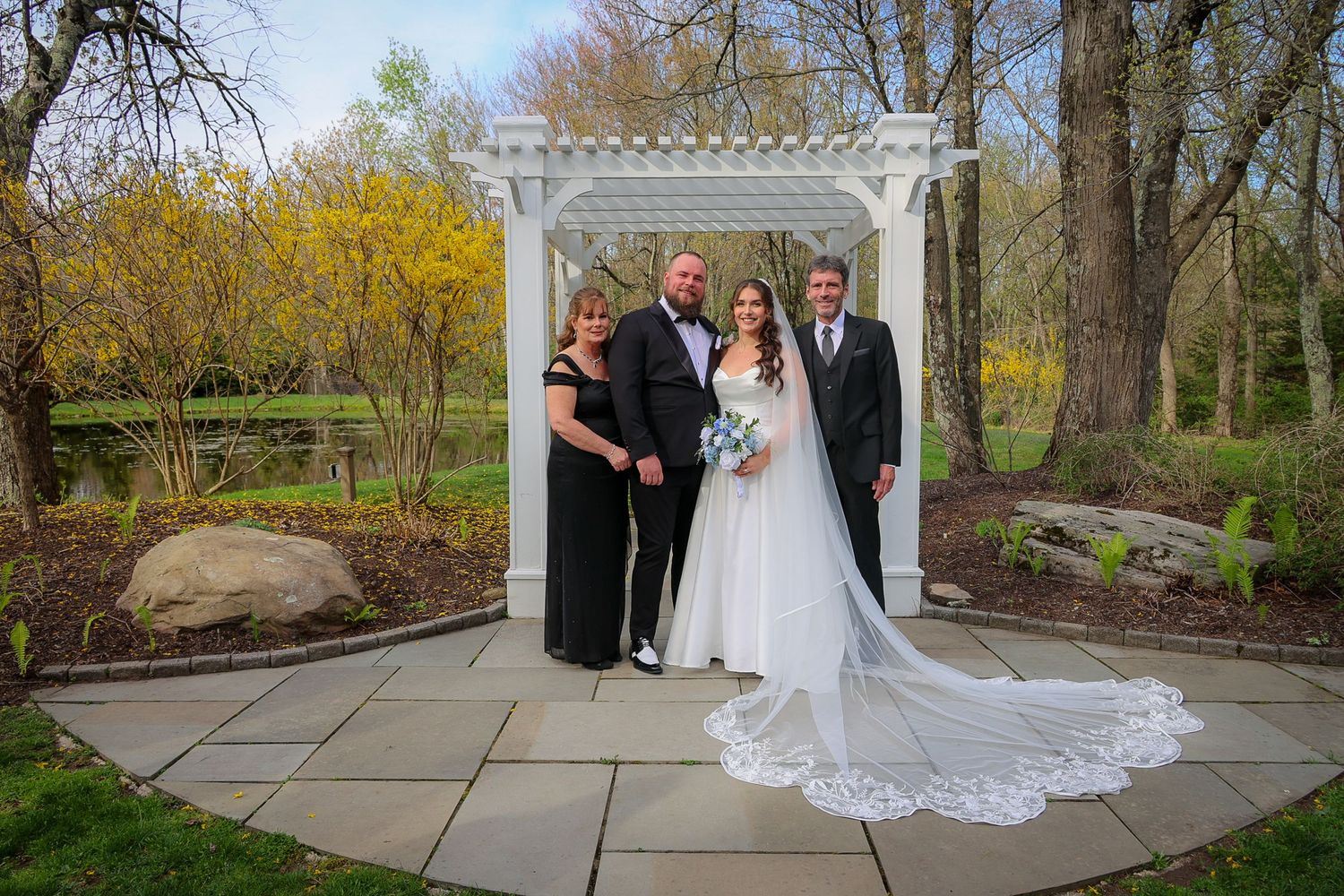Wedding party of four poses under white pergola in lush garden setting, bride in flowing gown with long veil.