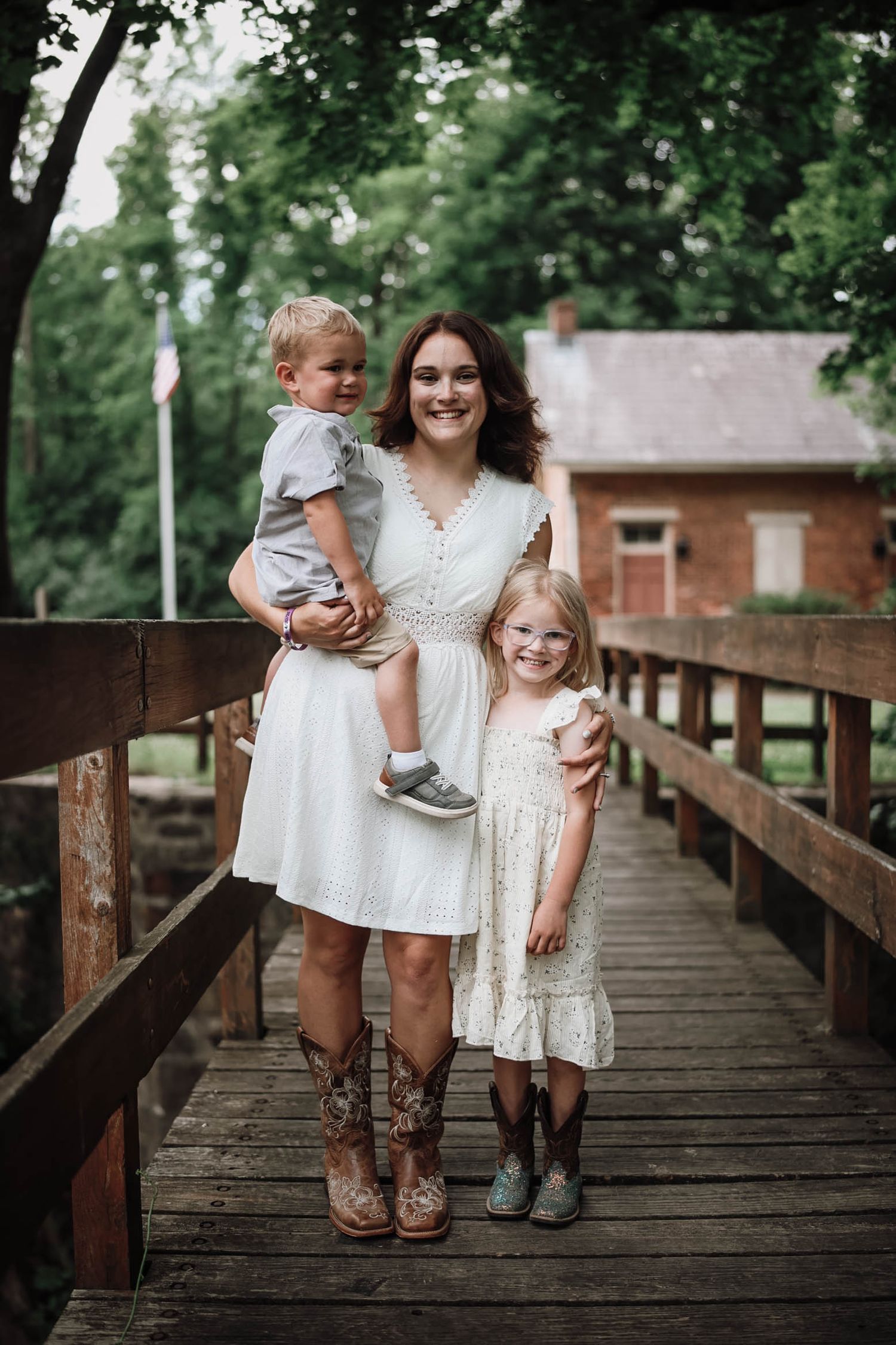 Mother in white dress and cowboy boots holding toddler with daughter on wooden bridge.