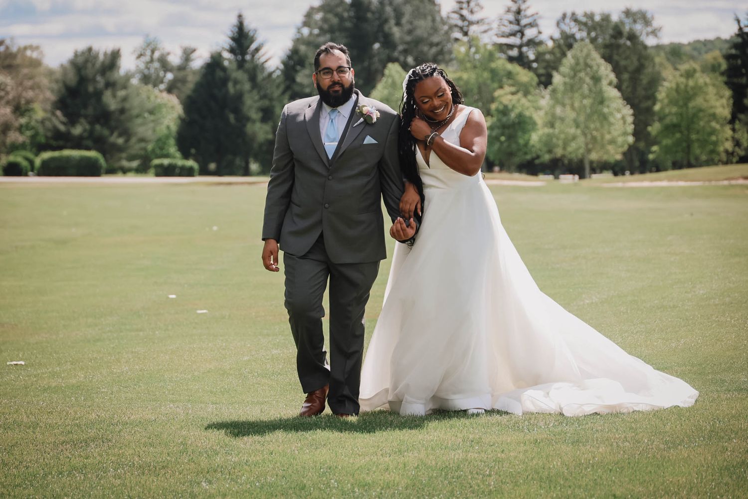 A bride in a white gown leans on her groom in a gray suit as they walk across a lush green lawn.