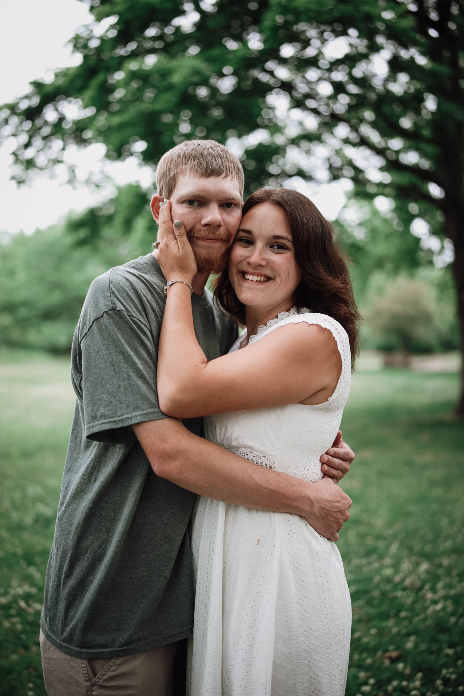 A smiling couple embraces outdoors, woman in white dress and man in gray tee, with lush green trees behind them.