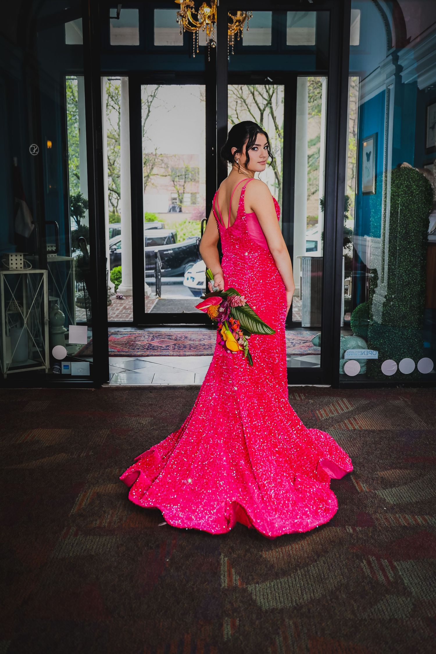 Woman in stunning hot pink sequin mermaid gown with train holds bouquet near elegant venue entrance doors.
