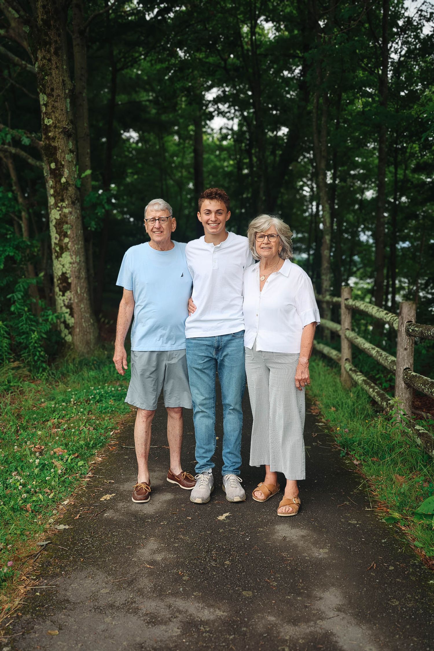 Three family members pose on a tree-lined path, with a young man standing between two smiling older adults.