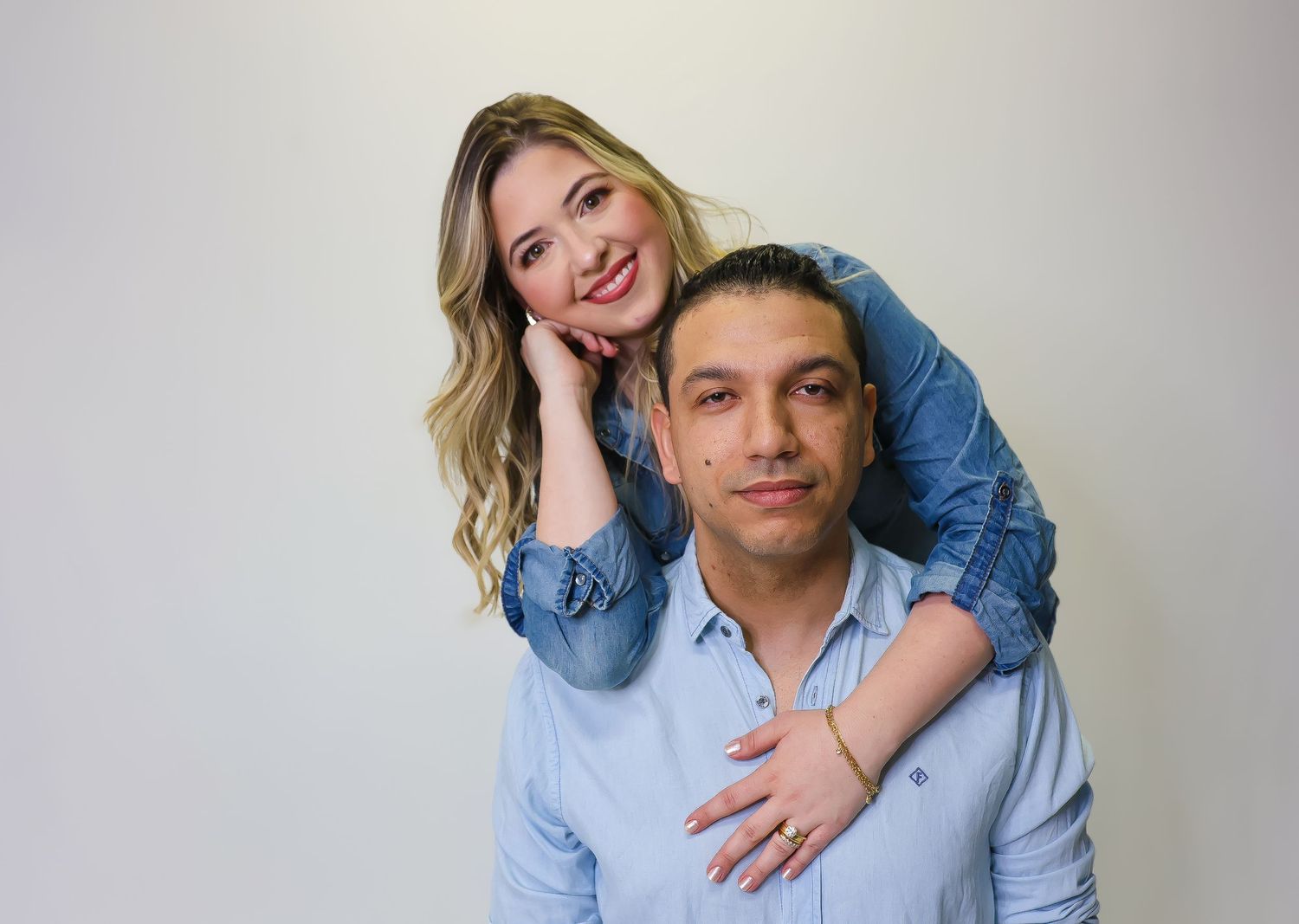 A smiling couple posing together, woman in denim jacket resting on man's shoulder against white background.