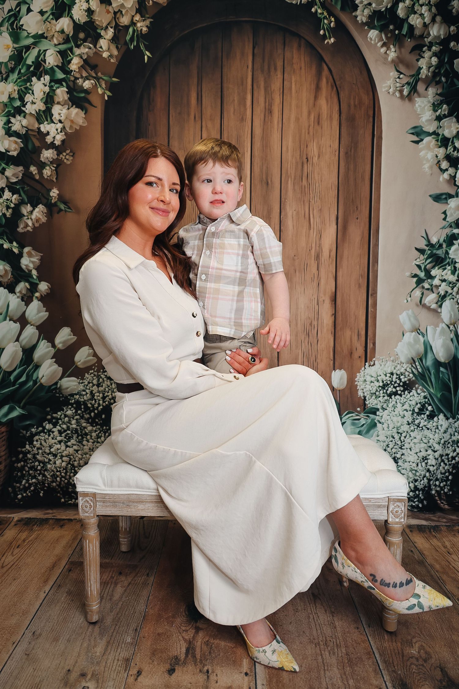 Woman in white dress sits with young boy in plaid shirt before floral arch backdrop with tulips.