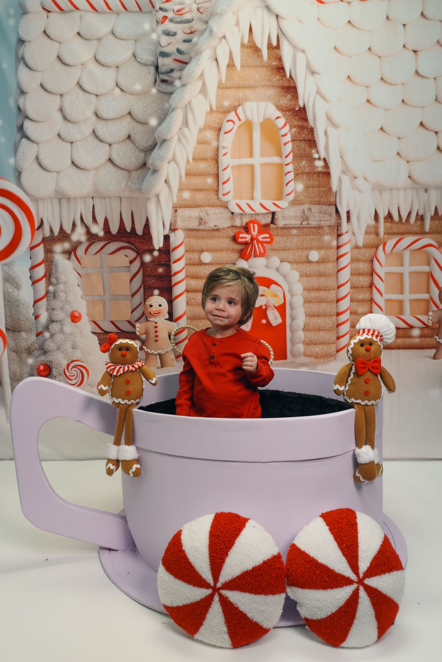 Smiling toddler in red outfit sits inside giant purple mug with gingerbread decorations against candy house backdrop.