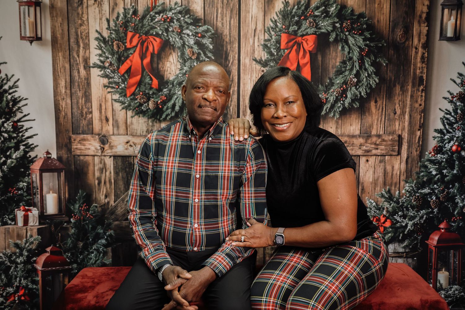 A couple smiles together in a festive Christmas photo session with holiday wreaths and plaid outfits.