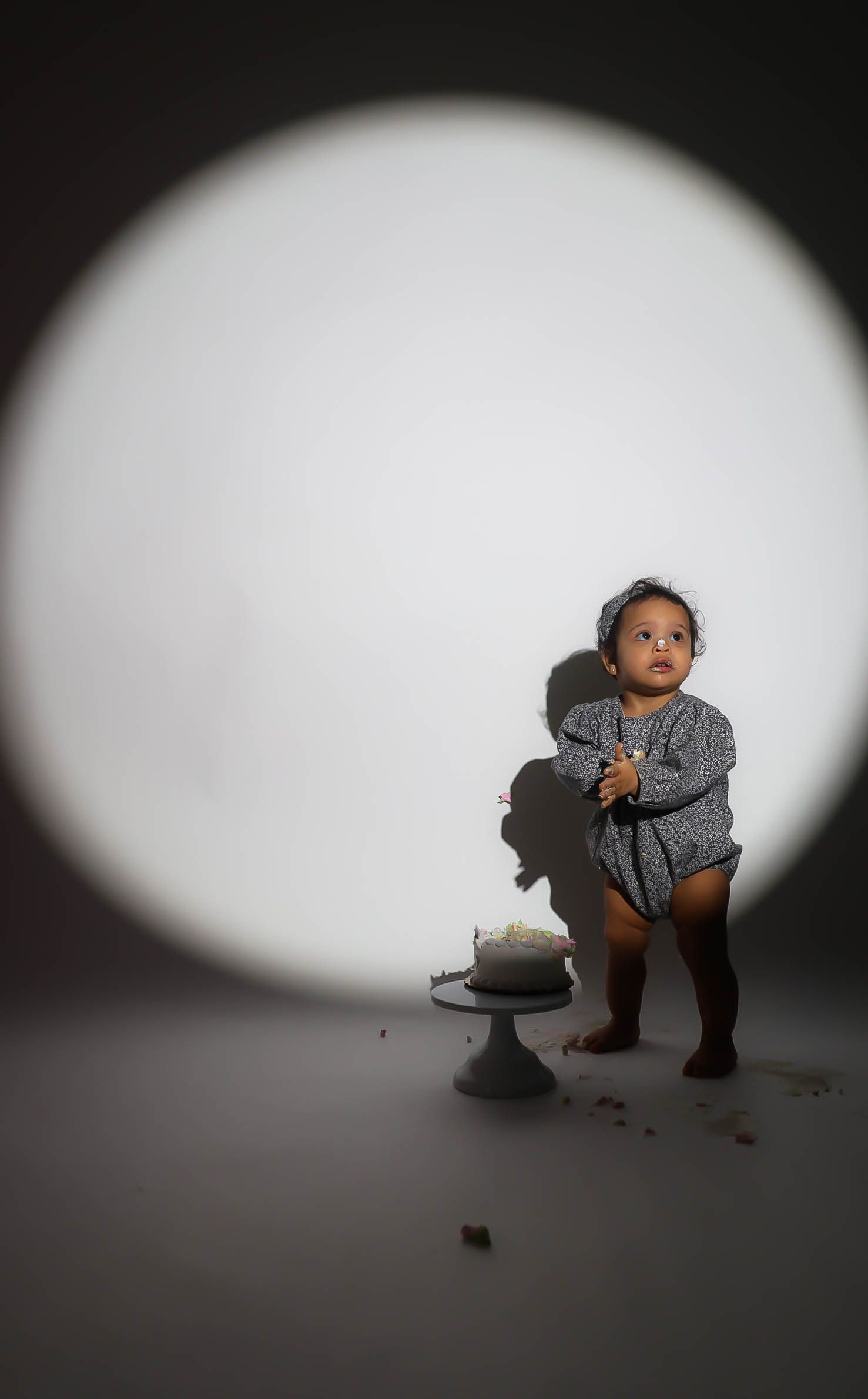 Baby in gray outfit stands by smashed first birthday cake in spotlight studio photo.
