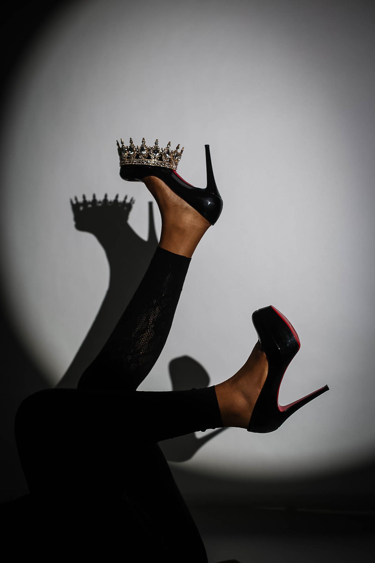 Legs in black heels balancing a silver crown under dramatic spotlight with shadow.