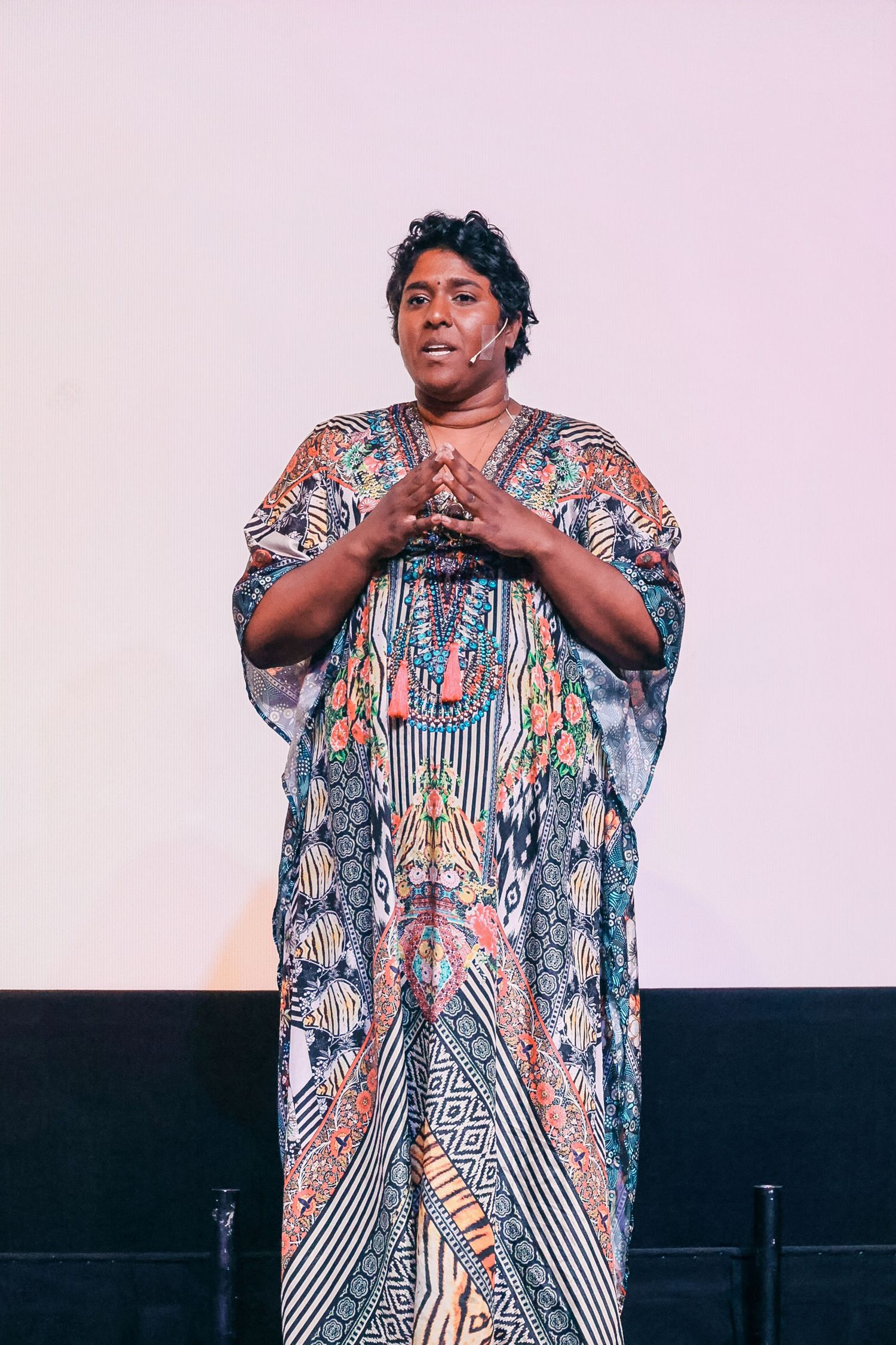 Woman in colorful patterned kaftan dress speaking on stage with hands clasped together.