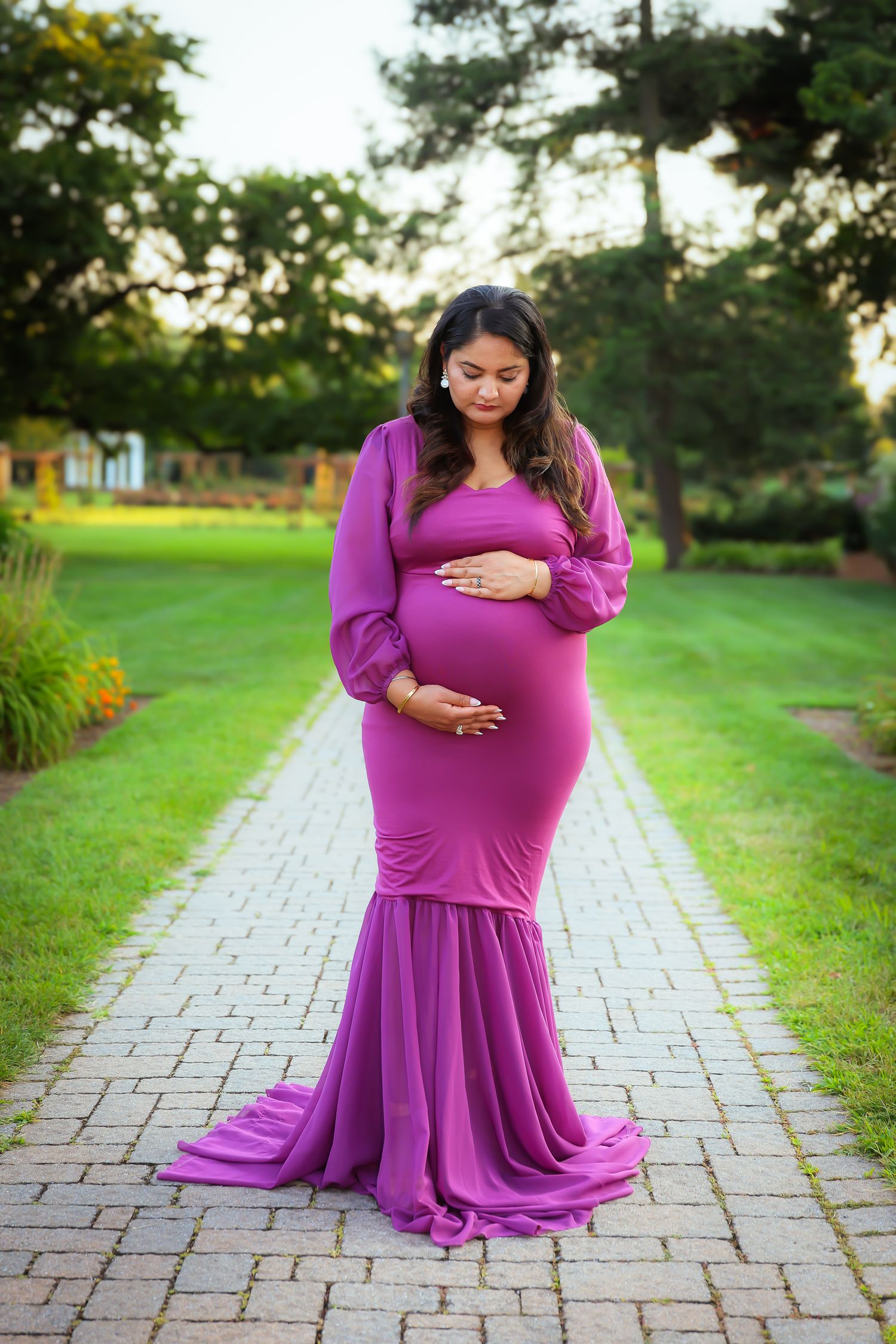 A woman in a flowing purple maternity dress stands on a brick pathway in a lush garden setting at sunset.