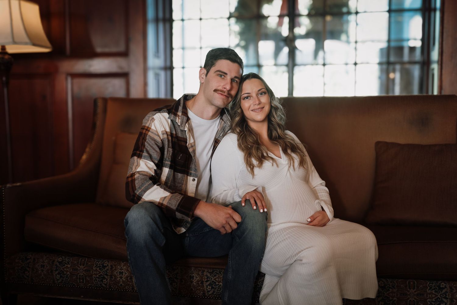 A couple in casual attire sits close together on a brown leather couch in a cozy indoor setting.